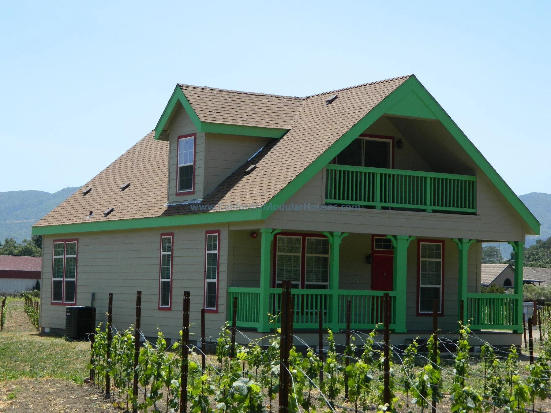 Two-story modular house with yellow siding and green trim, featuring a front porch, balcony, and mountain backdrop.