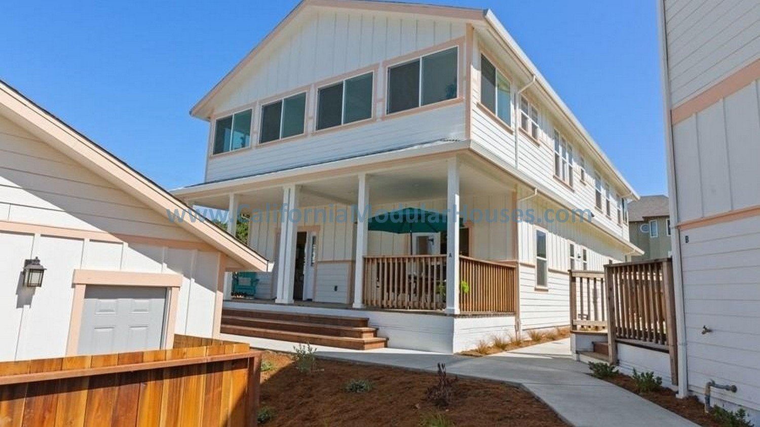 Newly prefab modular constructed white two-story house with a front porch & balcony , surrounded by a wooden fence and neighboring houses, under a clear blue sky.