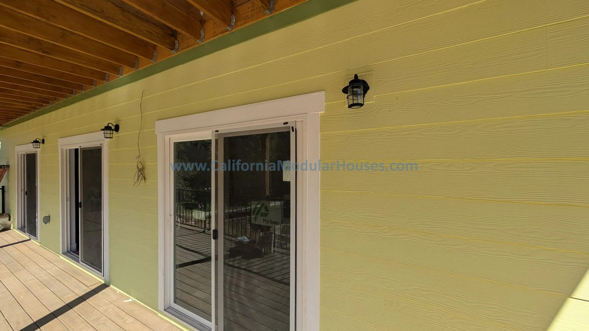 Yellow exterior wall of a house with three sliding glass doors, black outdoor wall lanterns, and a wooden deck.
