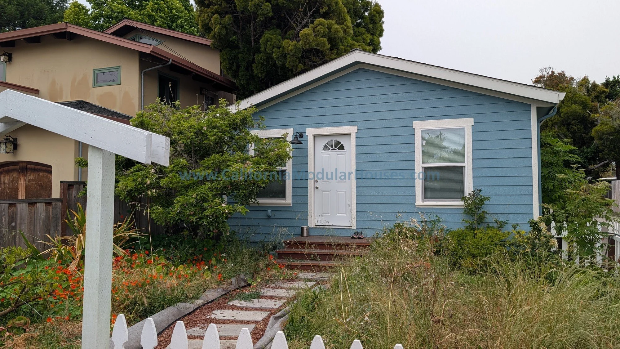 Front view of a small blue house with white trim, a white door, and two windows, surrounded by overgrown grass and plants, with a wooden staircase leading to the front door.