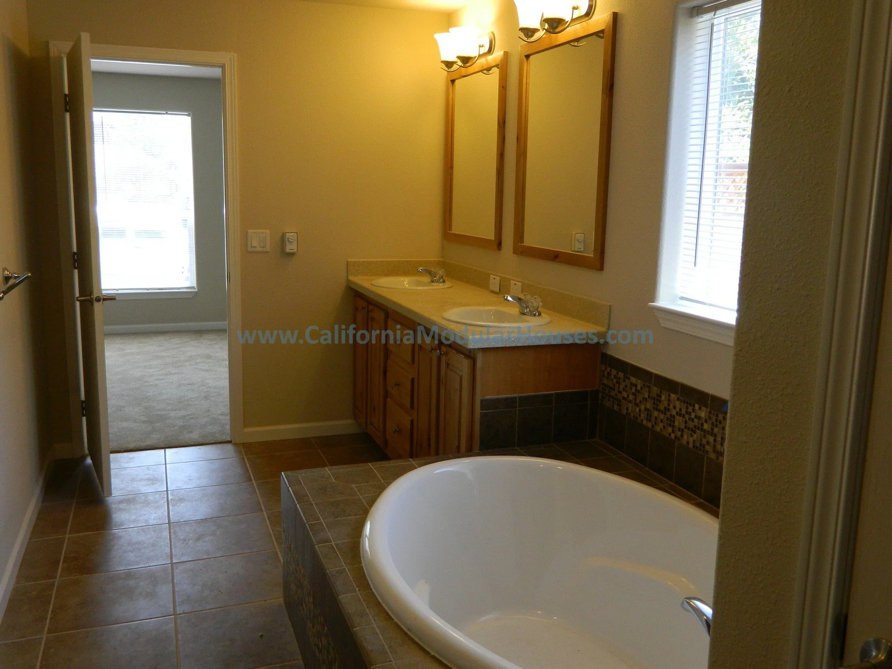 Bathroom with a corner bathtub, double sinks with wooden cabinets, mirrored medicine cabinets, tiled floor, and a window with blinds.