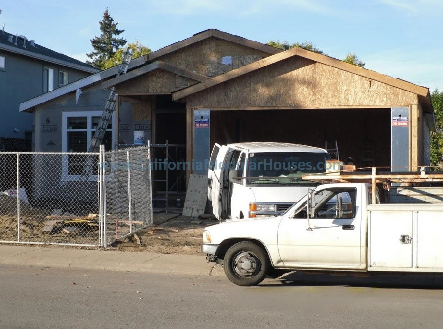 Under construction house with a partially built wooden structure, construction vehicles parked in front, and a chain-link fence surrounding the site.
