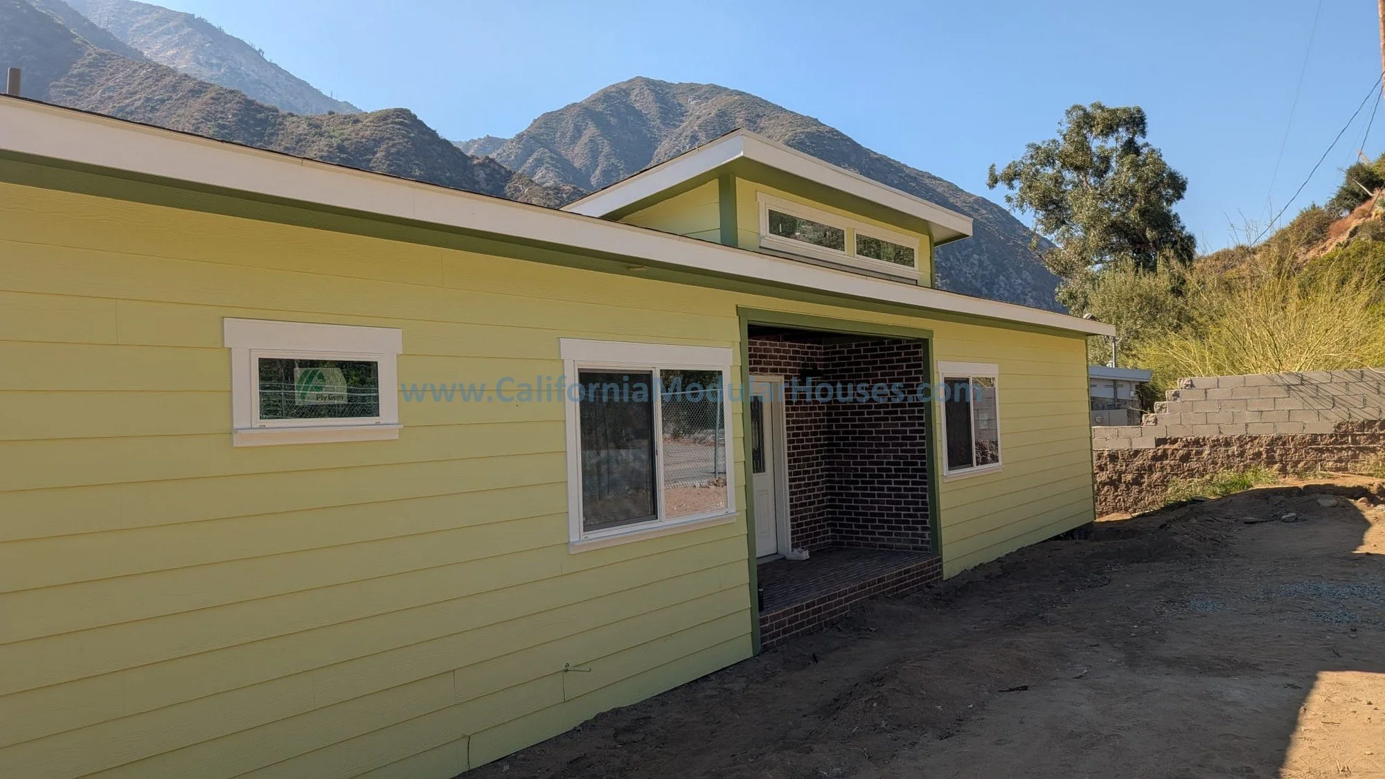Yellow house with white trim, three windows, a covered porch entry, and a mountainous background with trees and sunny sky.