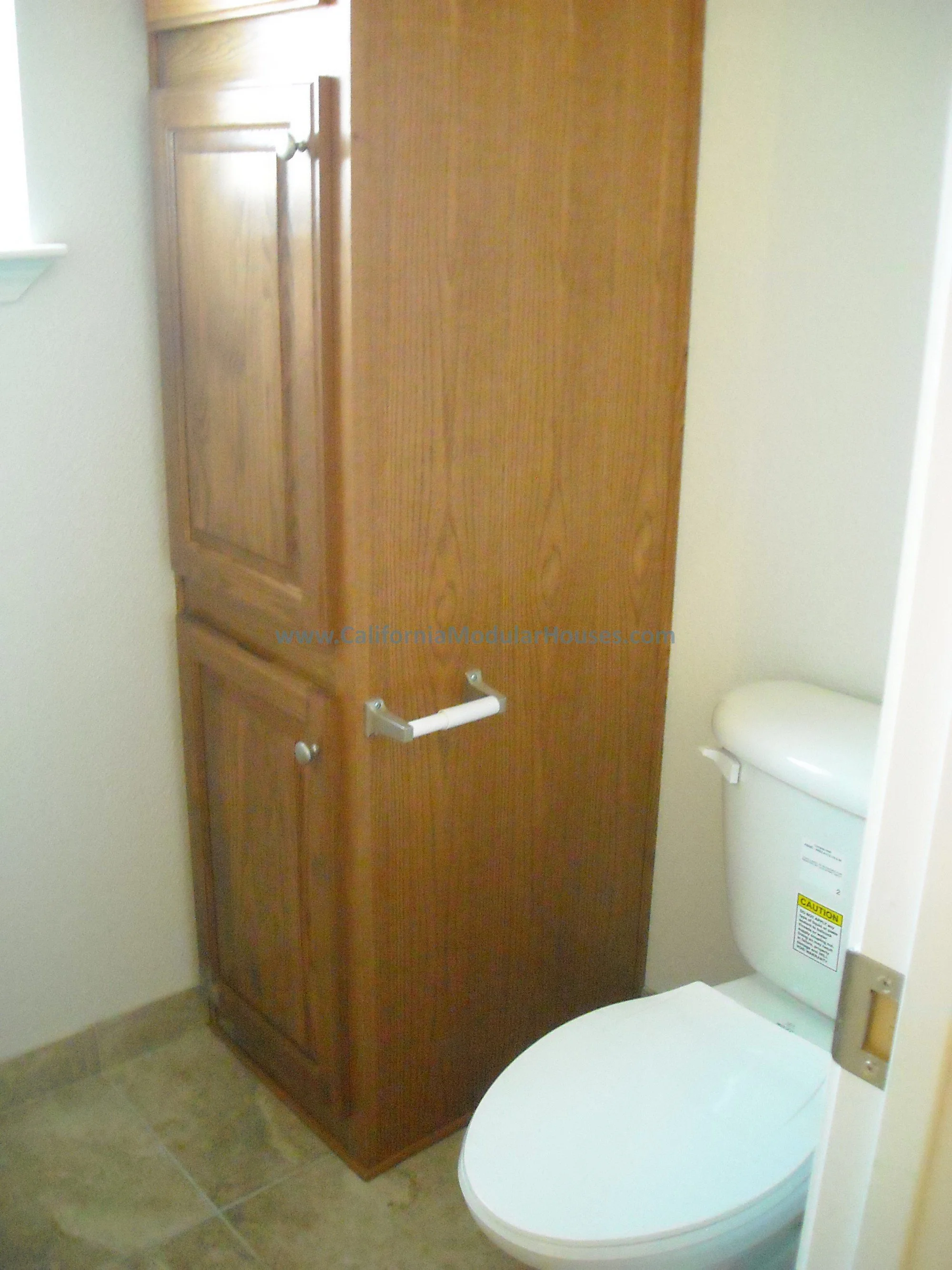A bathroom with a wooden cabinet beside a white toilet, beige tiled floor, and a small window on the wall.   This includes a linen cabinet in this bathroom for this prefab house.  
