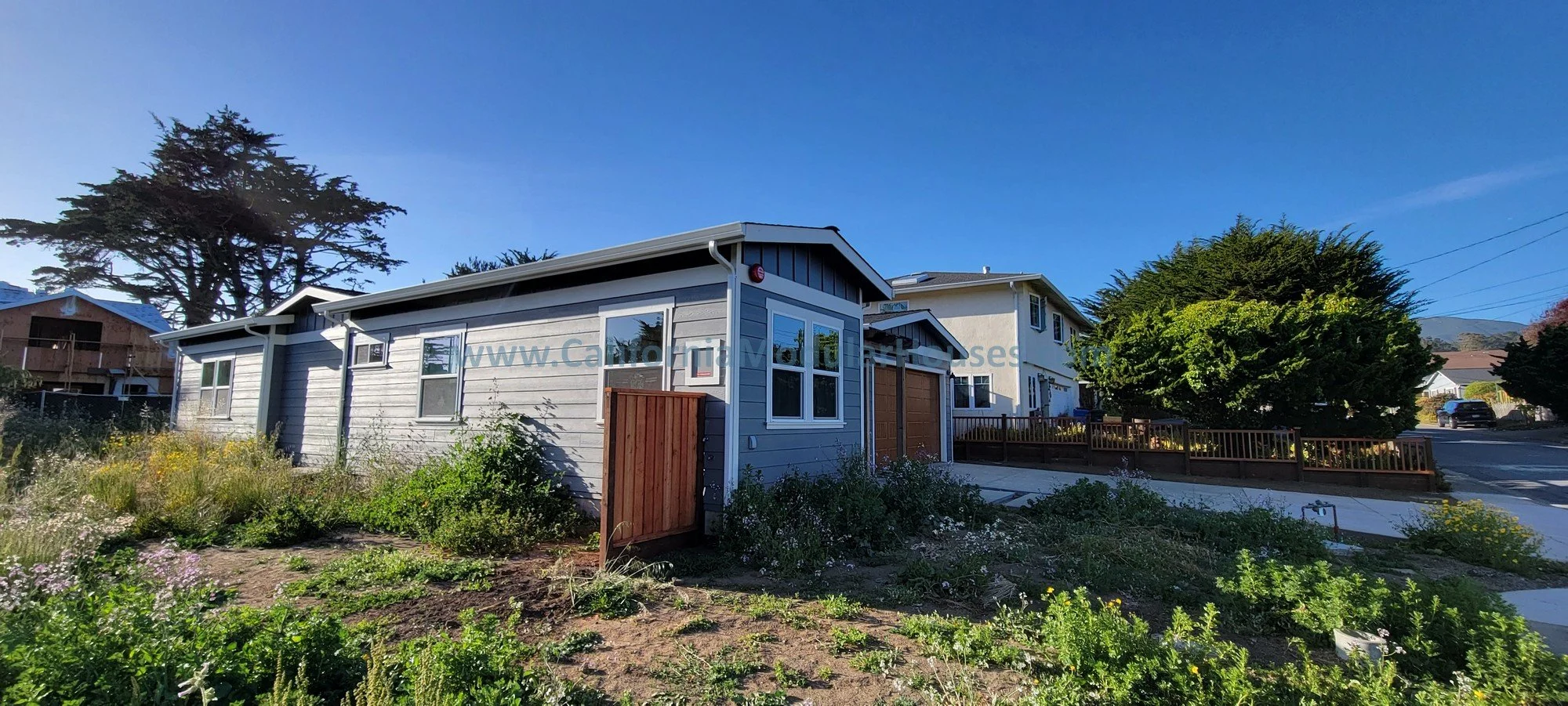 A residential neighborhood with a new light gray house in the foreground, a neighboring house in the background, greenery, trees, and a blue sky.