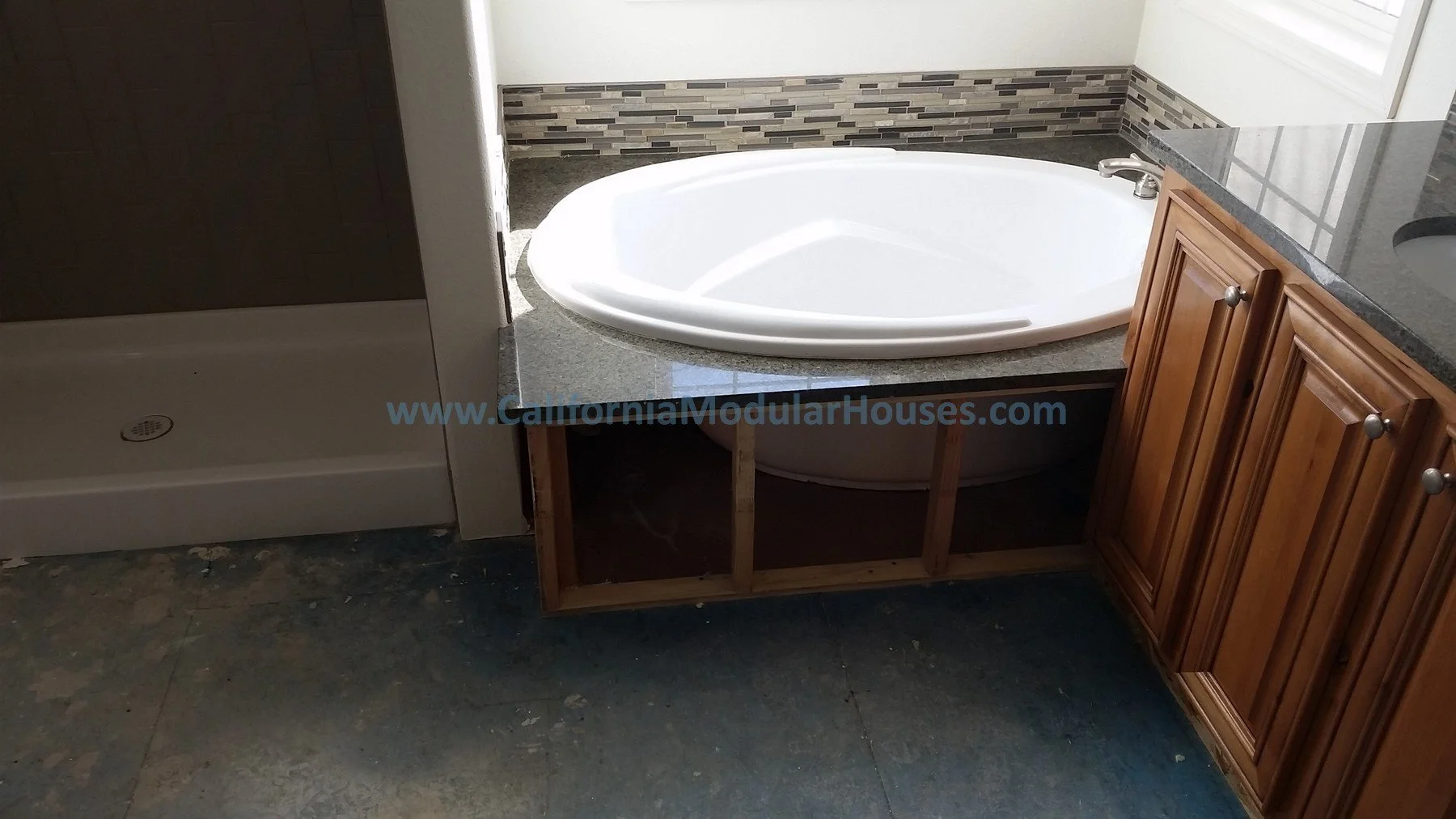Bathroom with a corner bathtub, wooden cabinet, granite countertop, and a shower stall to the left. The unfinished cabinet below the bathtub is visible. There is a window with sunlight coming in. Modular Home CA,