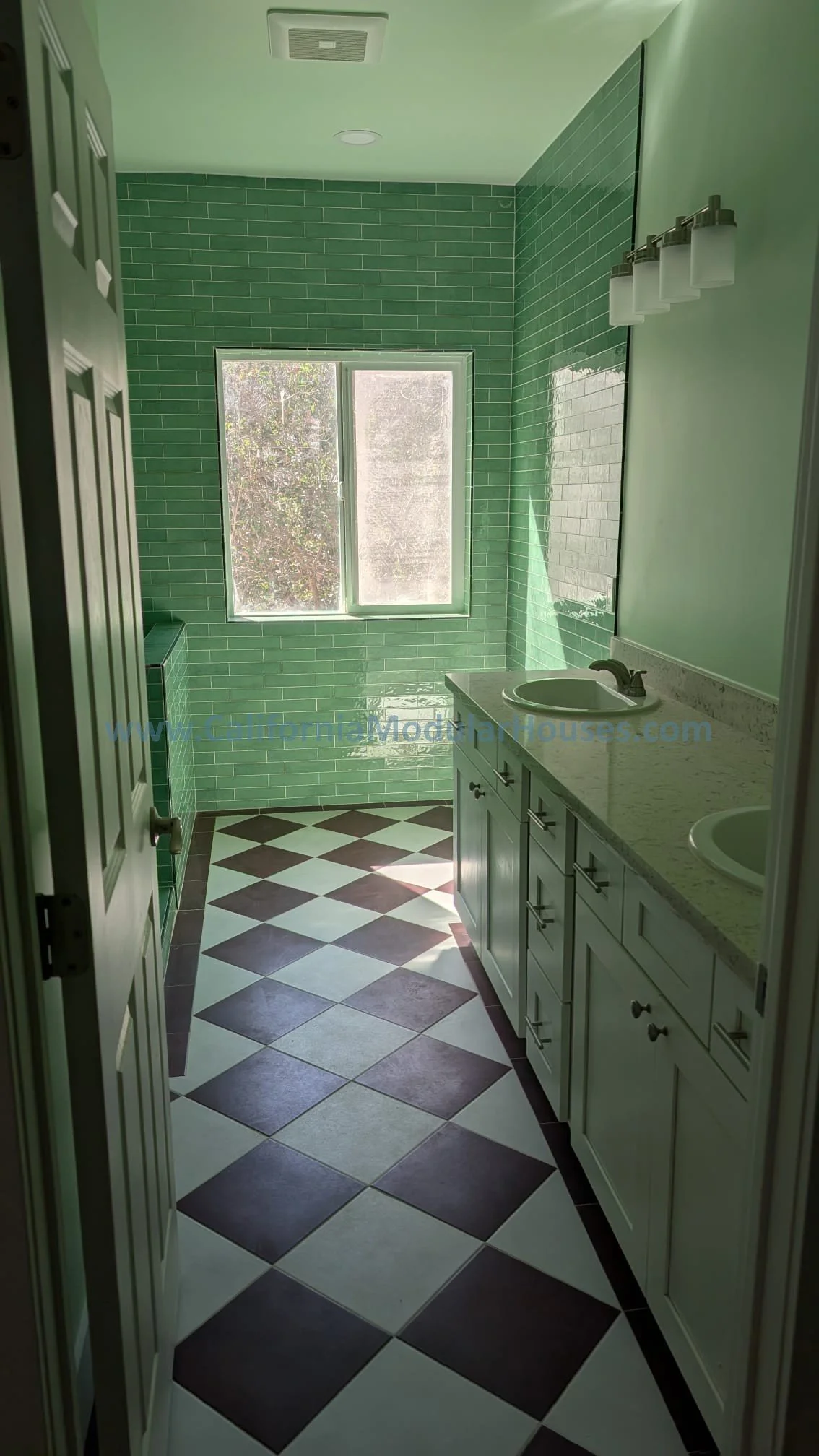 Bathroom with green tiled walls, double sinks with vanity, window, and black-and-white checkered floor.