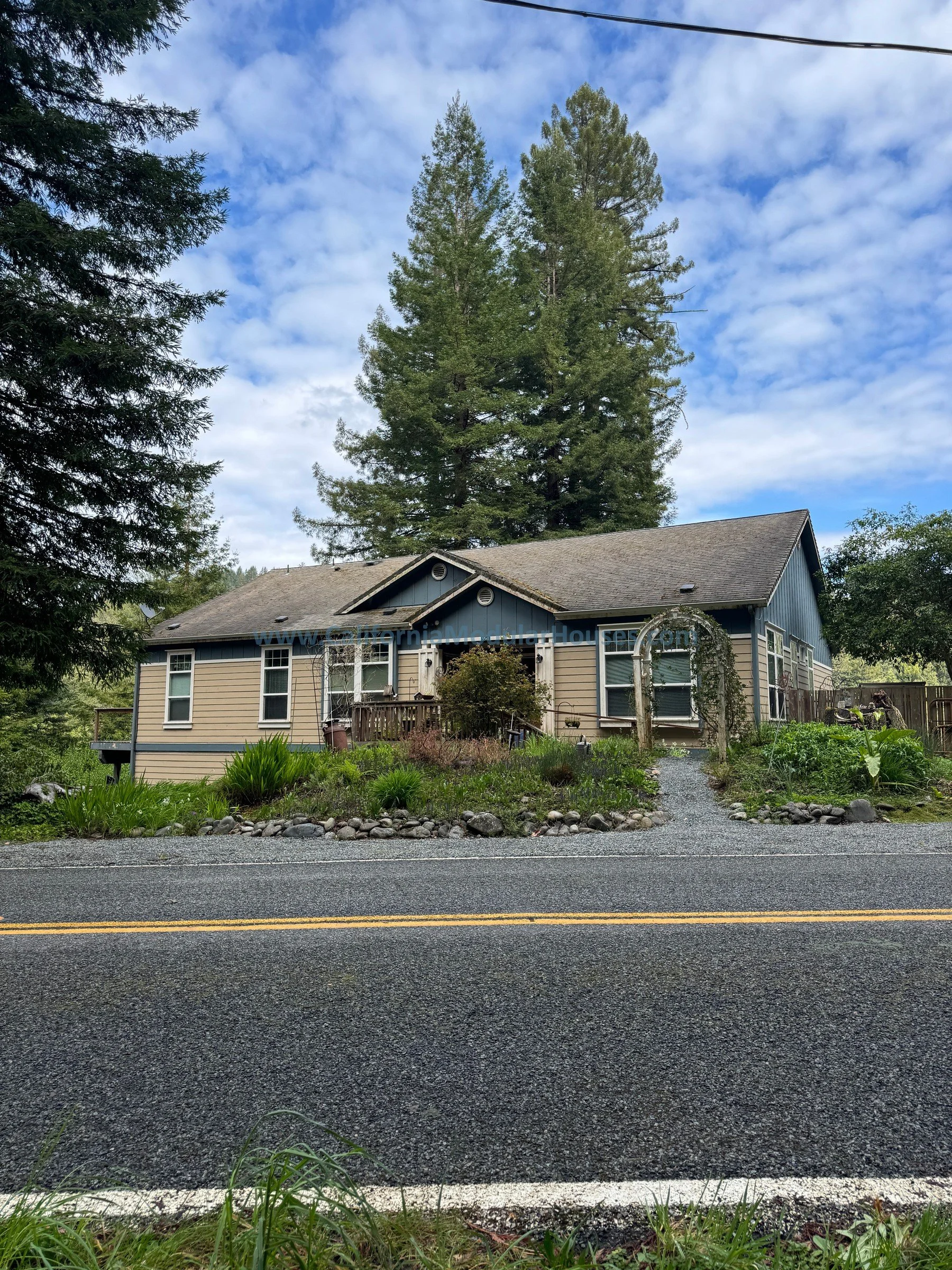 A house with beige siding and blue trim is situated behind a gravel driveway, surrounded by lush greenery and tall trees, under a partly cloudy sky. modular home california.  Crescent City, Del Norte County, CA.