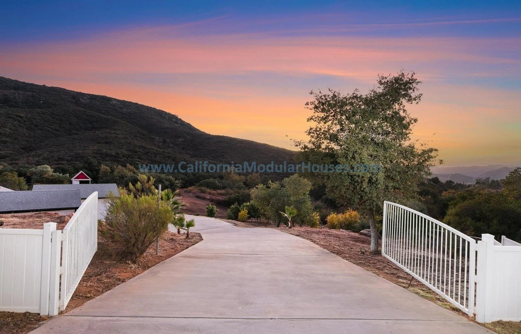 A scenic view at sunset with a winding concrete driveway, a white fence on both sides, a tree on the right, and rolling hills in the background under a colorful sky.