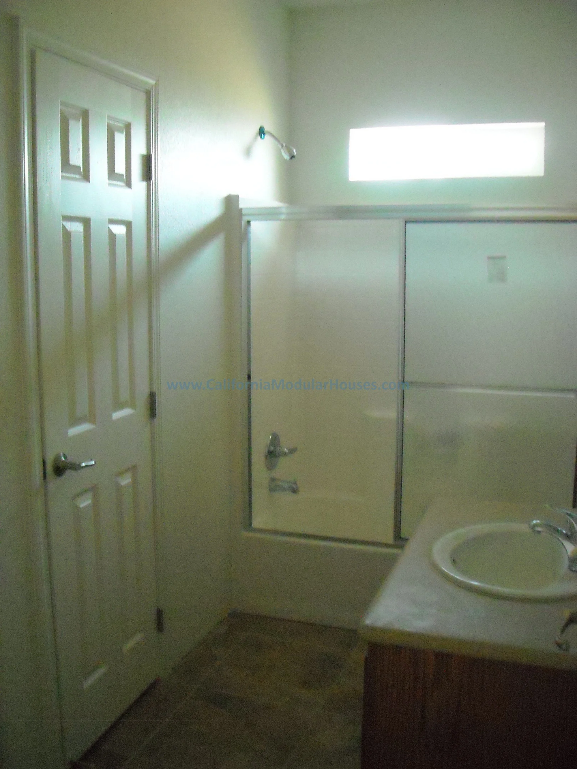 Photo of one bathroom with a closed six-panel door, a small frosted horizontal window, a shower with a glass door, a white sink with a faucet, and a wooden cabinet below the sink.  There is a composite tub/shower combo in this bathroom.  