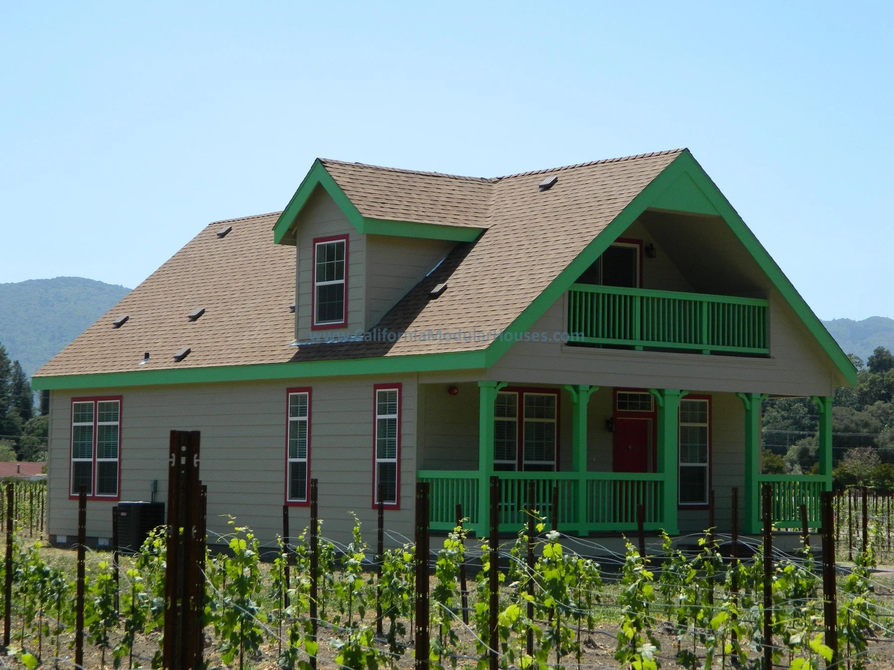 A two-story house with beige siding, green trim, and a gabled roof, set behind a vineyard with grapevines.