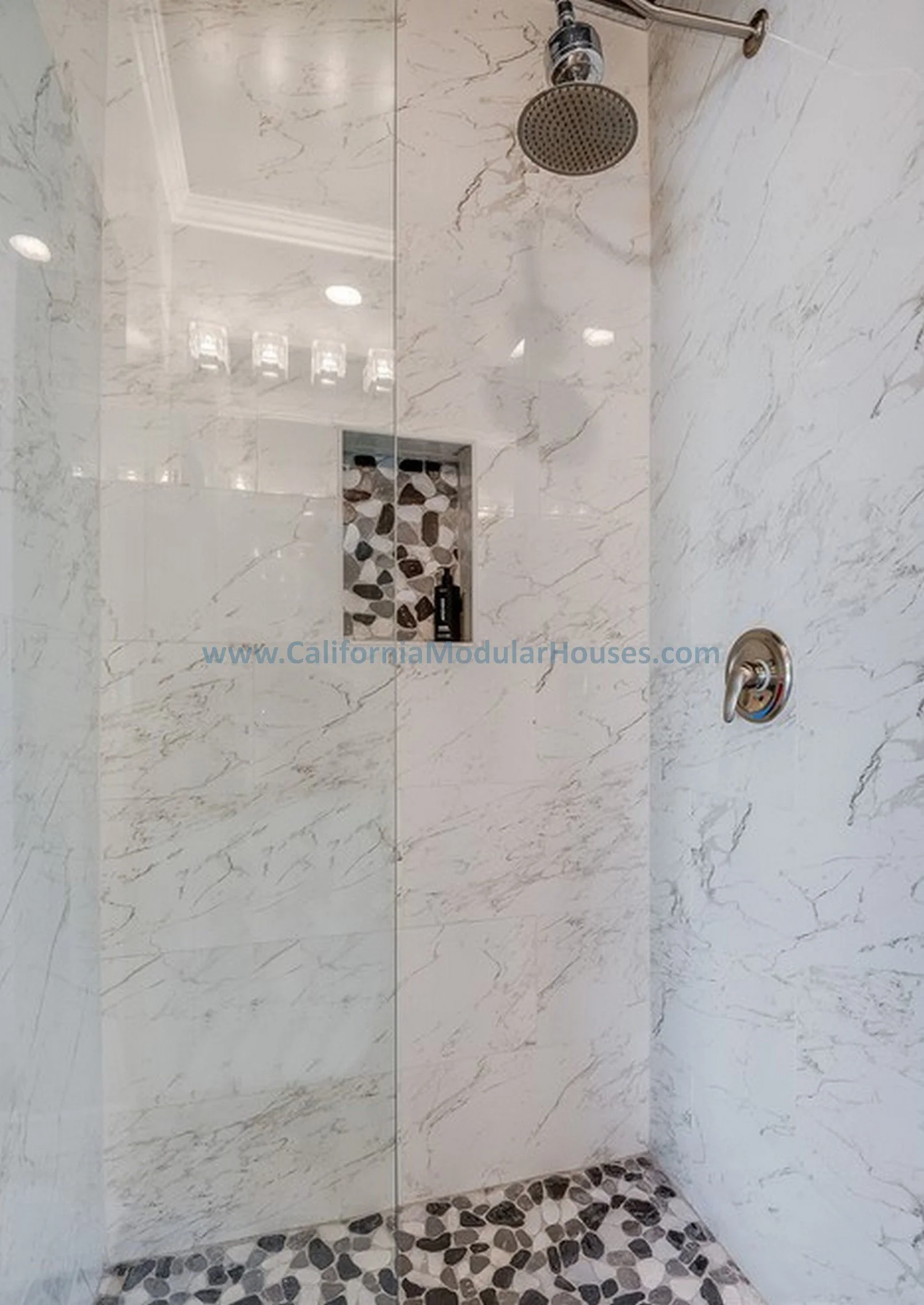Modern shower with white marble walls, a rainfall showerhead, and a pebble tile floor. There is a recessed niche with decorative stones and a bottle of shampoo inside.  Factory Built Housing in Southern California.  