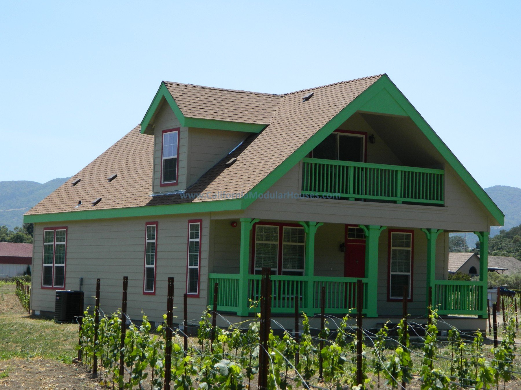 A two-story house with beige siding, green trim, and a brown shingled roof. The house has a front porch and balcony, surrounded by a garden with rows of green plants and a clear blue sky in the background.