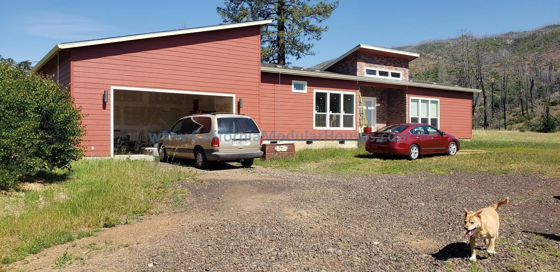 A modern house with a red exterior, large windows, and a garage.  The front yard, which has a mix of grass and gravel, with mountain scenery in the background.  California Modular, California Modular Home.