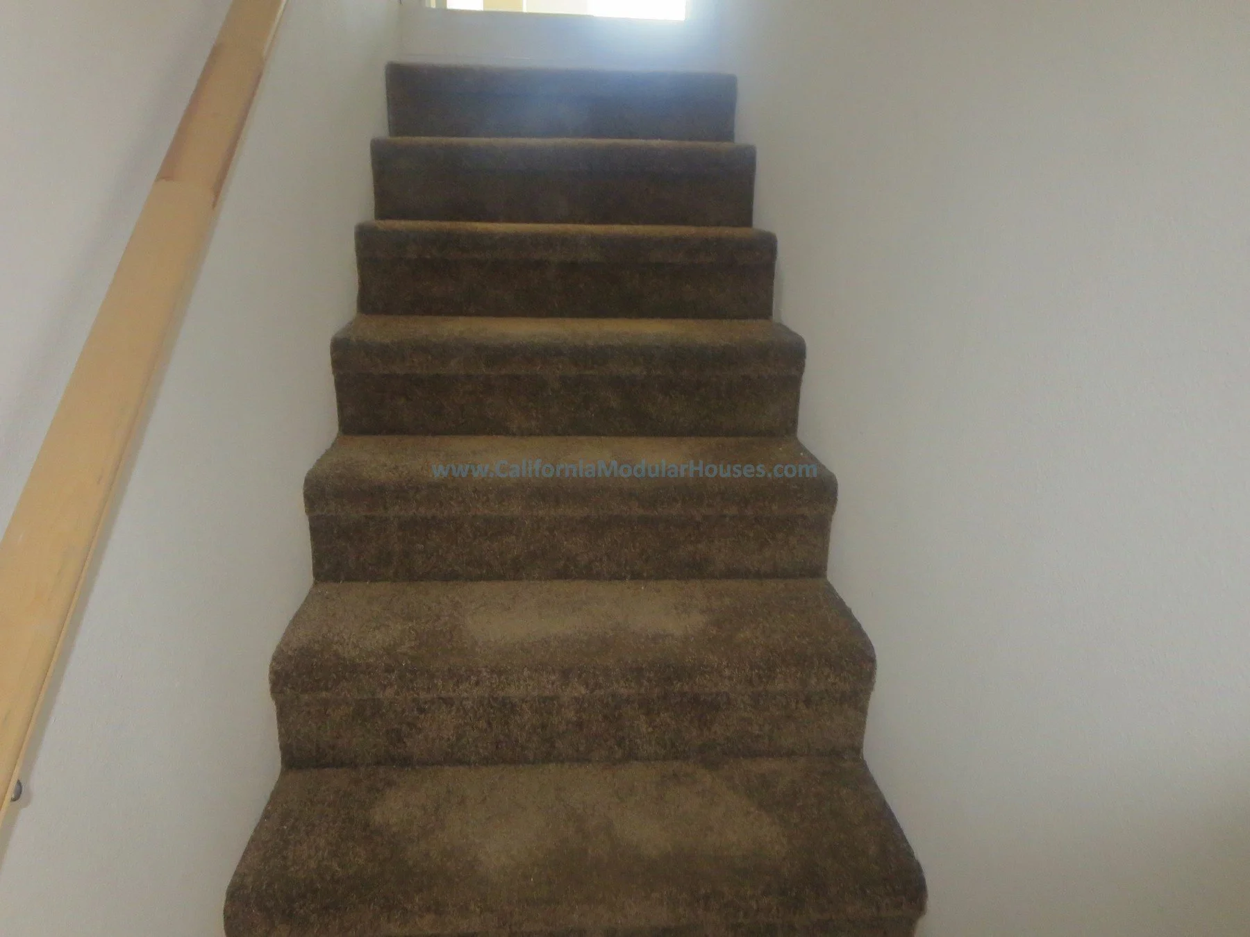 Indoor staircase with brown carpet, white wall on the right, wooden handrail on the left, leading upwards towards a source of bright light.  Unfinished attic.  New prefab modular house in Napa, CA.  California modular home.  
