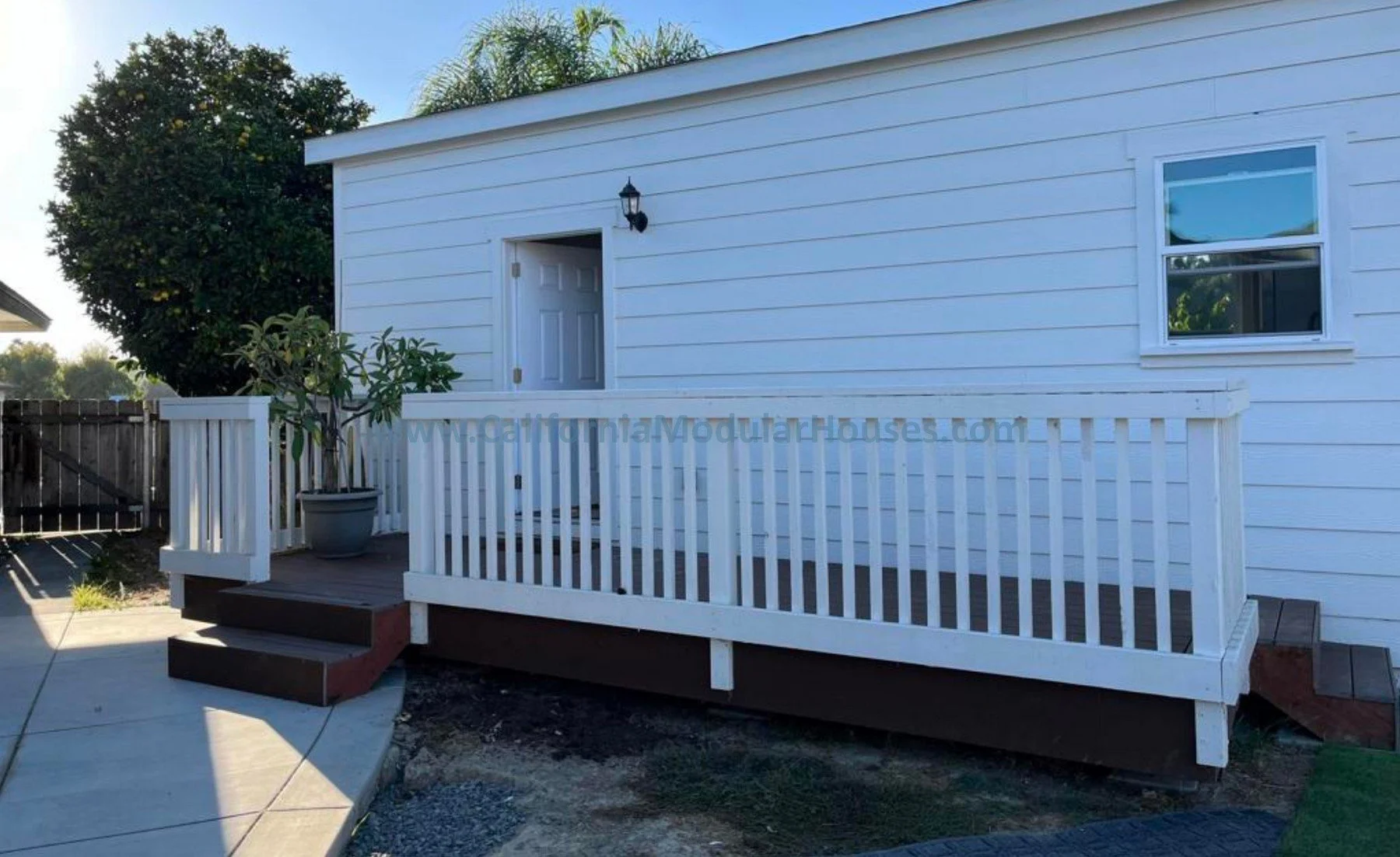 Backyard porch with white railing, steps leading up to a door, potted plant, and a wall-mounted outdoor light, with a white house exterior and a large tree in the background.