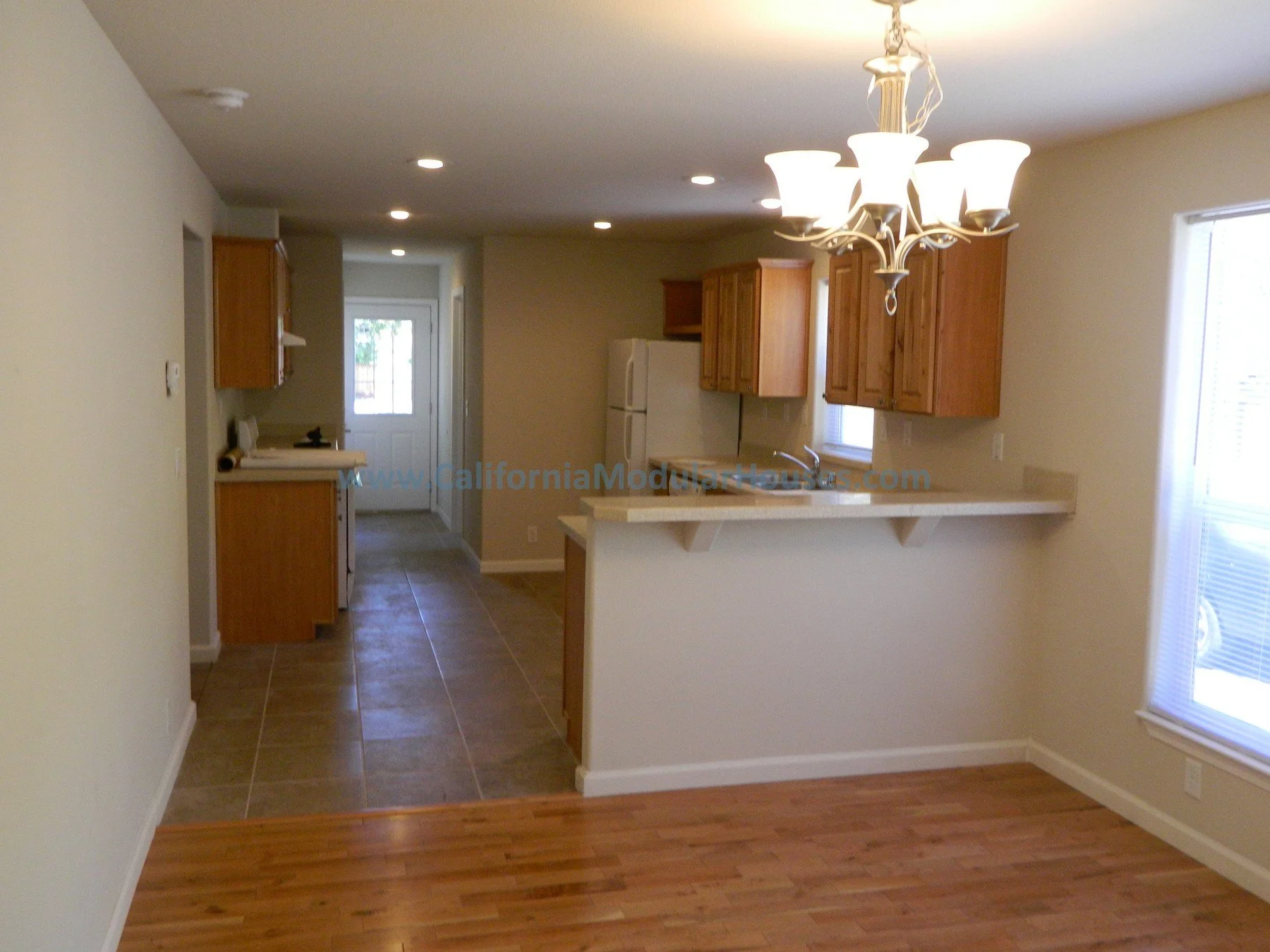 Kitchen area with wooden cabinets, a white refrigerator, a small breakfast bar, and a chandelier hanging over the dining space, natural light from a window, and flooring transitioning from hardwood to tile.