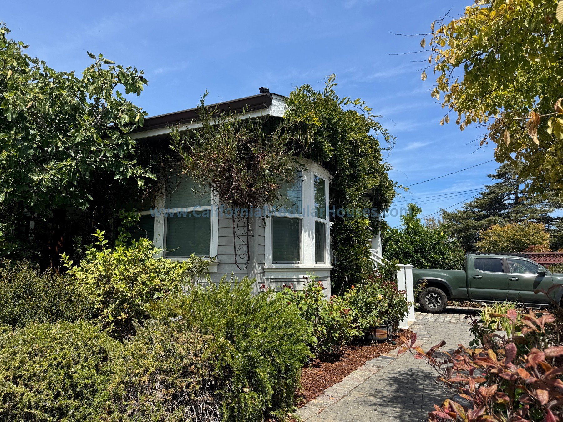 A white house with large windows, overgrown plants on the roof, surrounded by a garden with lush greenery, parking area with a parked dark green pickup truck, and a stone-paved driveway during daylight with a clear blue sky.