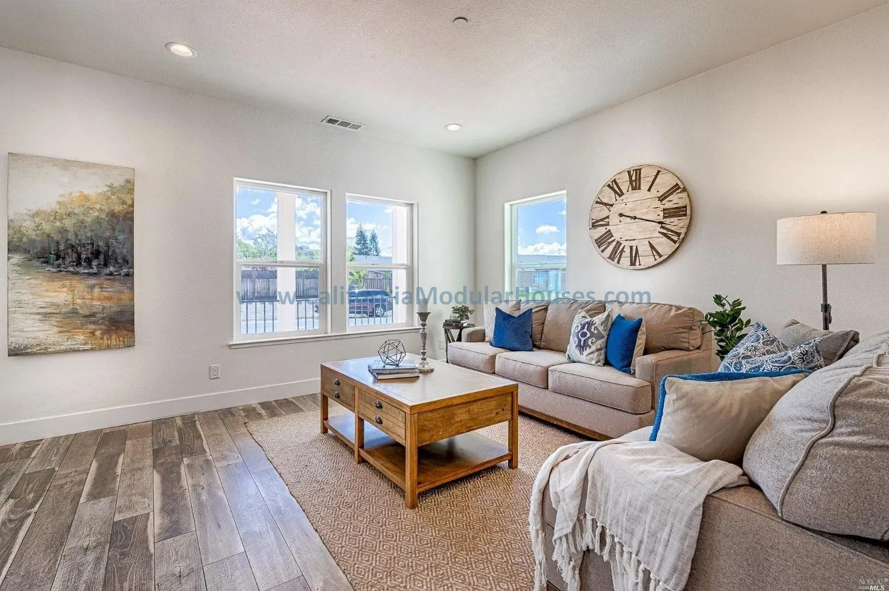 Living room with beige sofas, blue pillows, a wooden coffee table, a large wall clock, framed art, window views of a fence and trees, and a floor lamp.