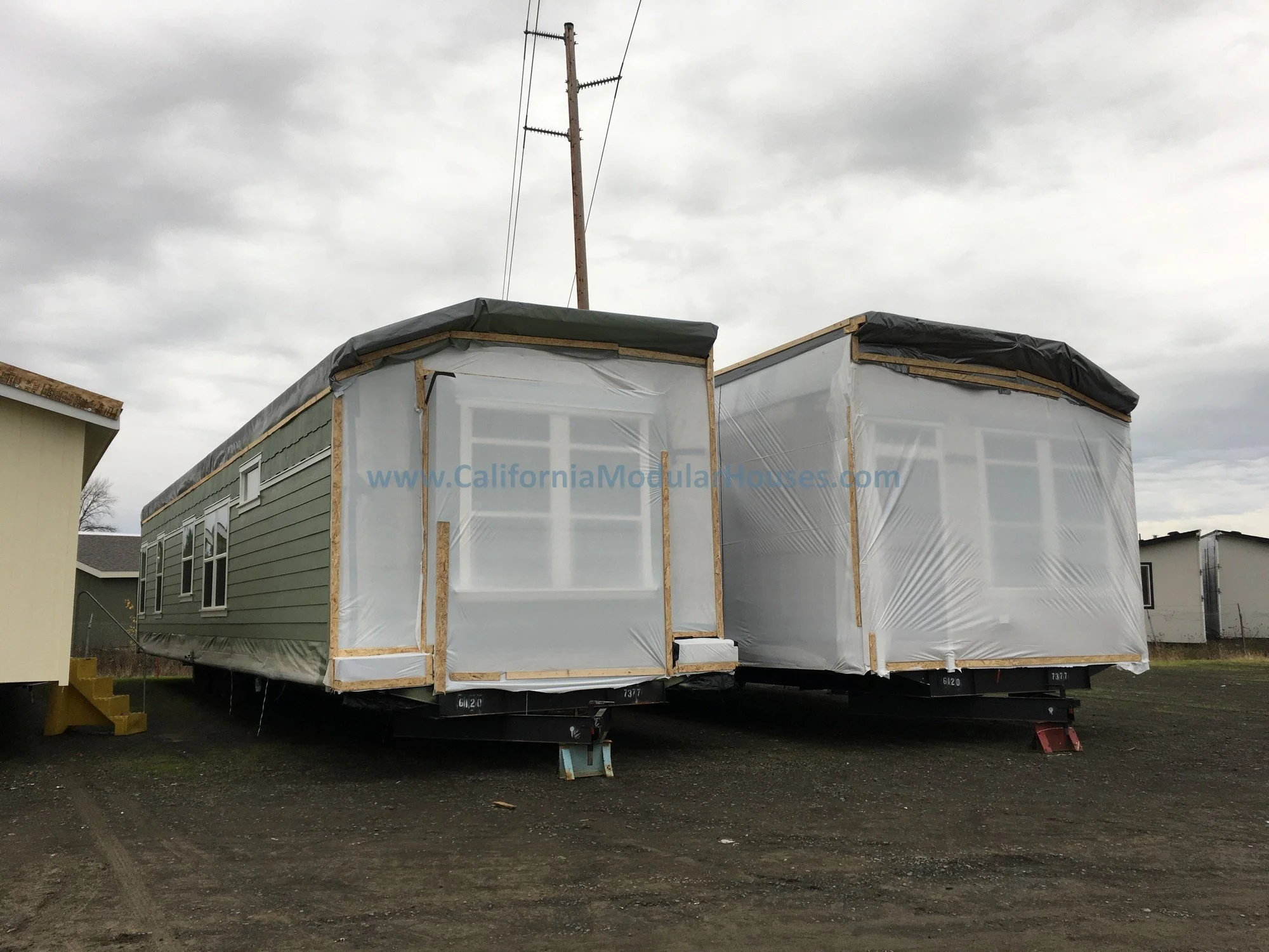 Two prefab modular homes under construction on a dirt lot, wrapped in white plastic and black roofing material, with neighboring houses visible in the background.