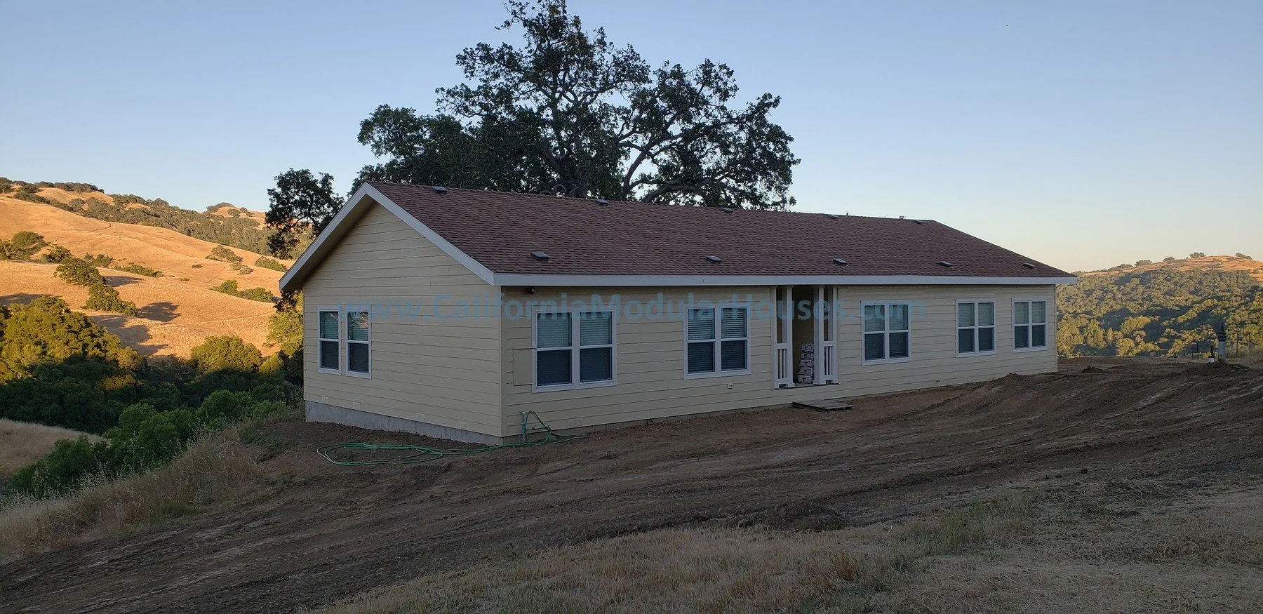 A newly constructed, light yellow house with white trim and multiple windows, situated on a hillside with a landscape of rolling hills and trees in the background during daytime.  Bay Area Prefab Homes, Modular Home CA.