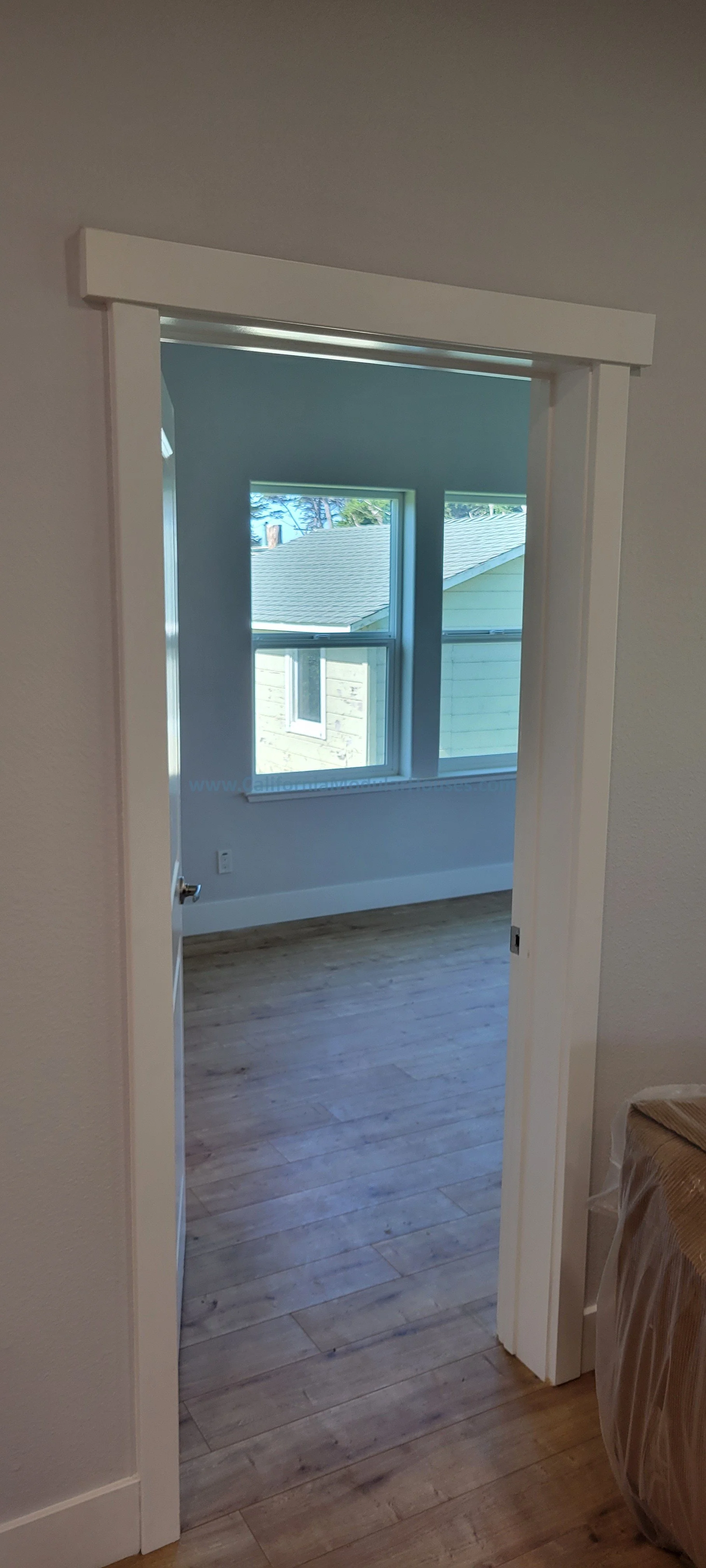 View through a doorway into a room with large windows, light blue walls, and wooden flooring.
