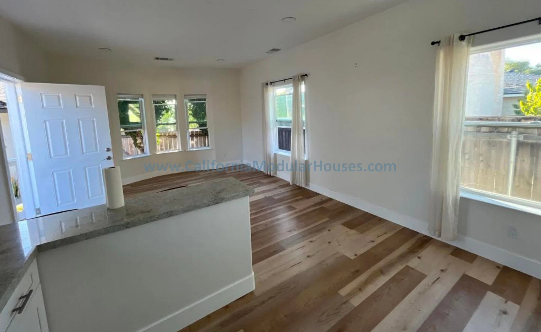 Empty living room with hardwood floors, multiple windows with curtains, and an open door showing outdoor fencing.  Interior view of prefab modular home.  Modular Home CA.  Oceanside, San Diego County, CA.  