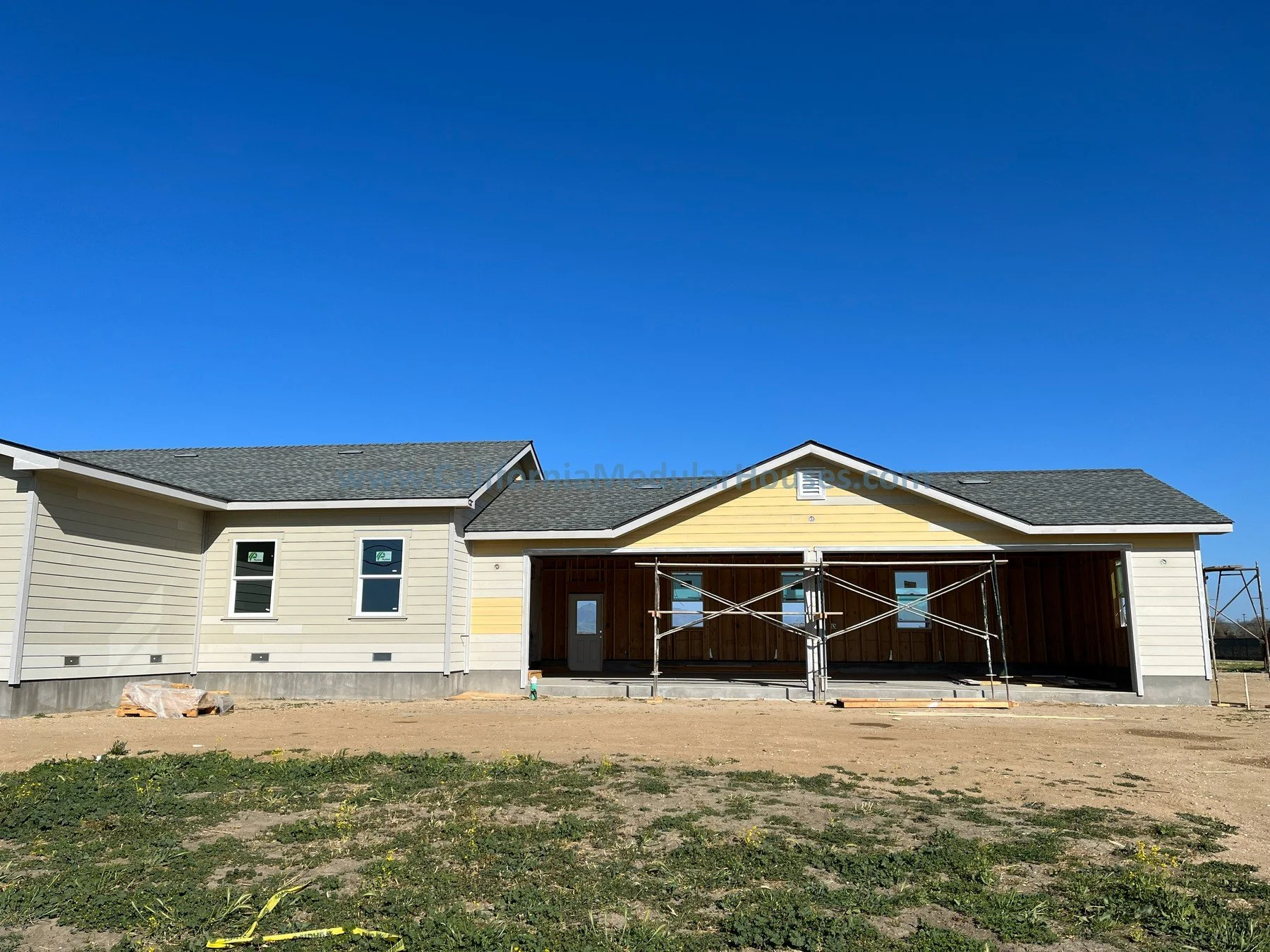 A modular house under construction with beige and yellow siding, a gray shingled roof, and a porch area with scaffolding. Garage is nearing completion.  California