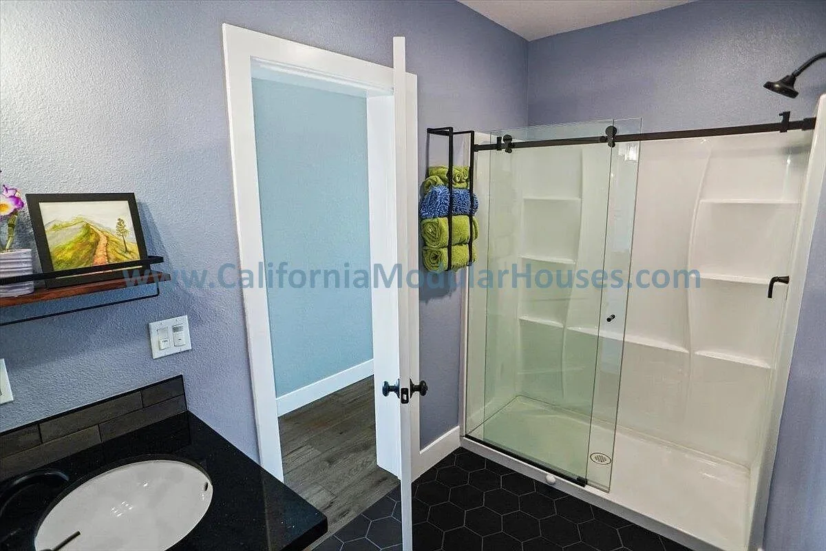 Bathroom with a corner shower with sliding glass doors, white shower walls with built-in shelves, a black countertop with an oval sink, a floating shelf with a framed landscape painting, purple-painted walls, and a doorway to a room with blue walls.