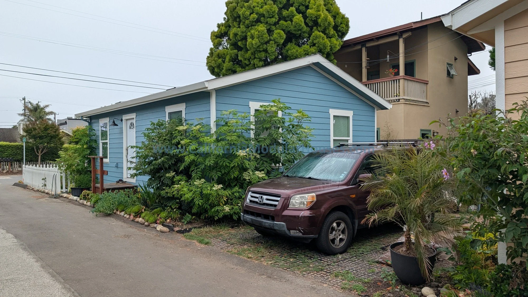 A small blue house with white trim, surrounded by greenery and flowers, with a maroon Honda Pilot parked in front, and two neighboring buildings visible in the background on a cloudy day.