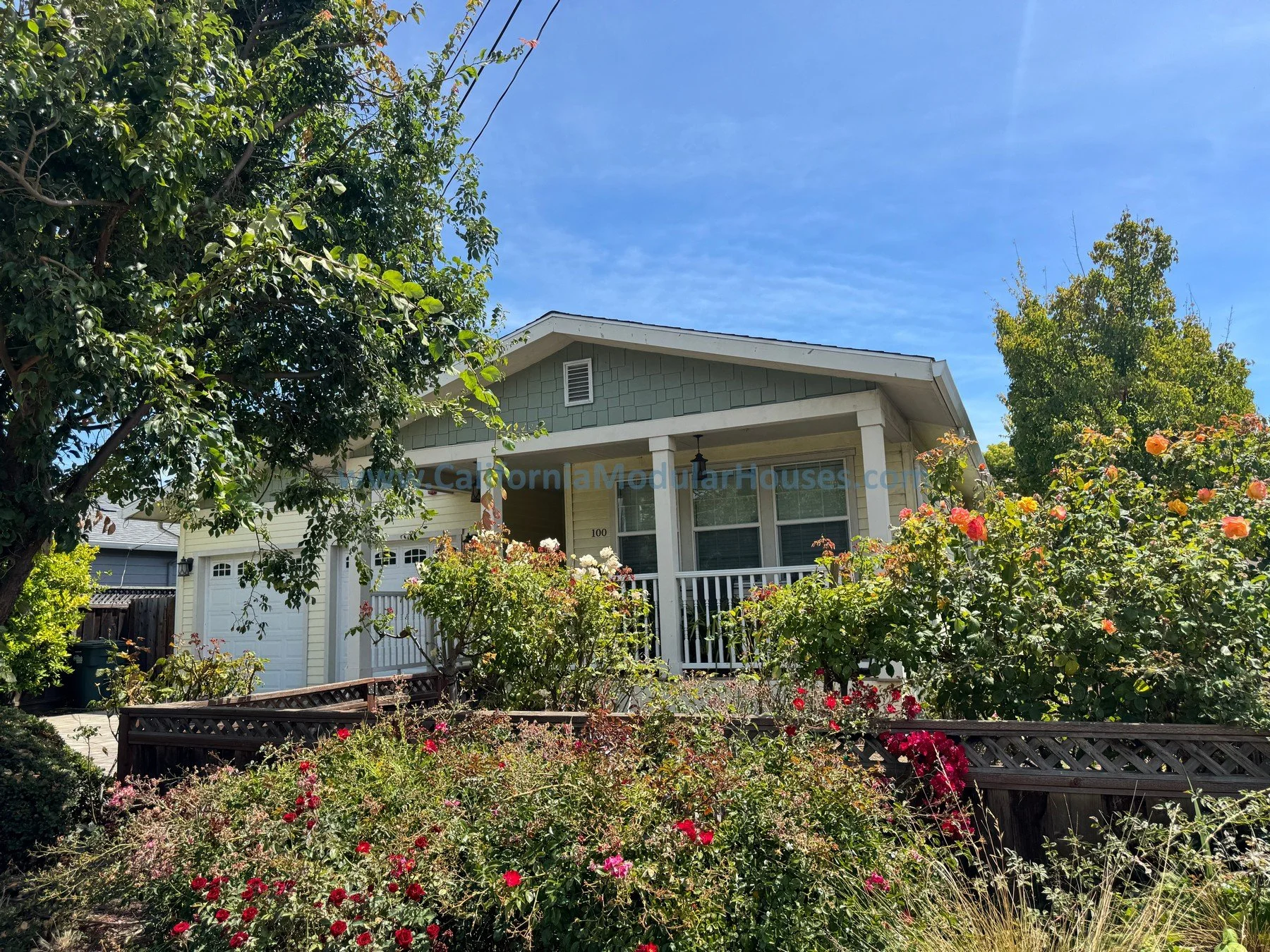 A two-story house with light green siding, white trim, a front porch, and a white garage door, surrounded by a lush garden with blooming roses and trees under a blue sky.