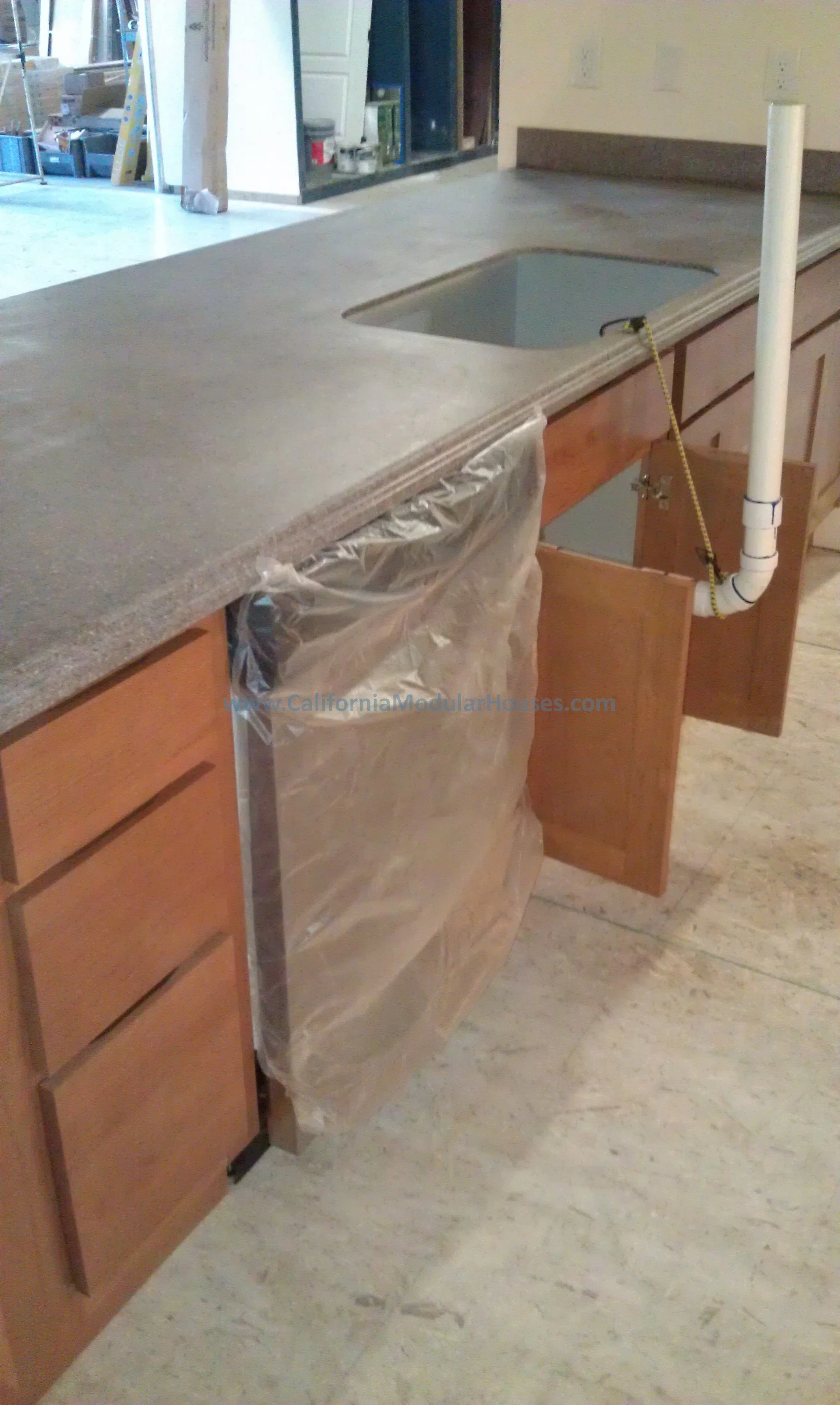 Kitchen countertop under construction with plumbing in place for a sink, cabinets beneath, and a plastic covering on one cabinet door. Prefab Home California, Pre-Fabricated Homes.