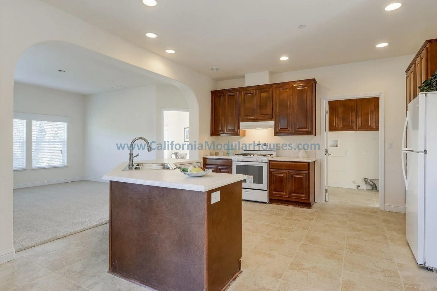 View of a kitchen with beige tiled floors, white walls, wooden cabinets, white appliances, and an island with a stainless steel sink.  Pre-Fabricated Homes, Modular Homes in California, Modular Homes California,