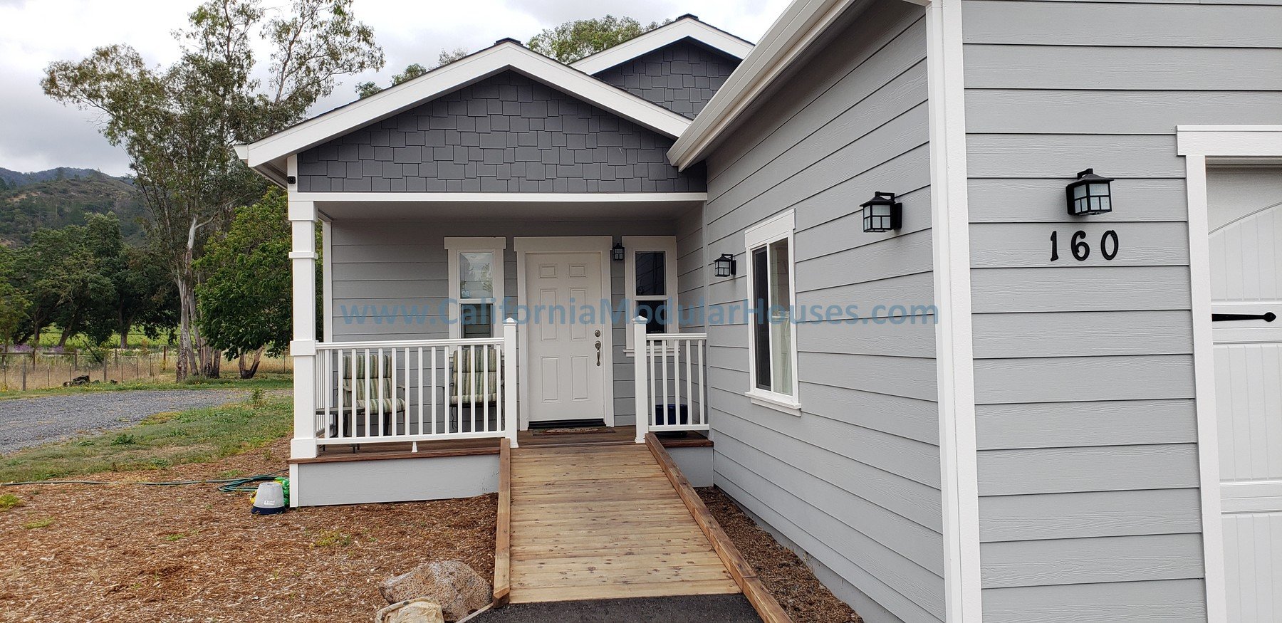 Front exterior of a gray house with a white door, a small porch with a wooden ramp, and black wall-mounted lights.  Prefab modular ADU with attached single car garage.   Kenwood, Sonoma County, CA. 