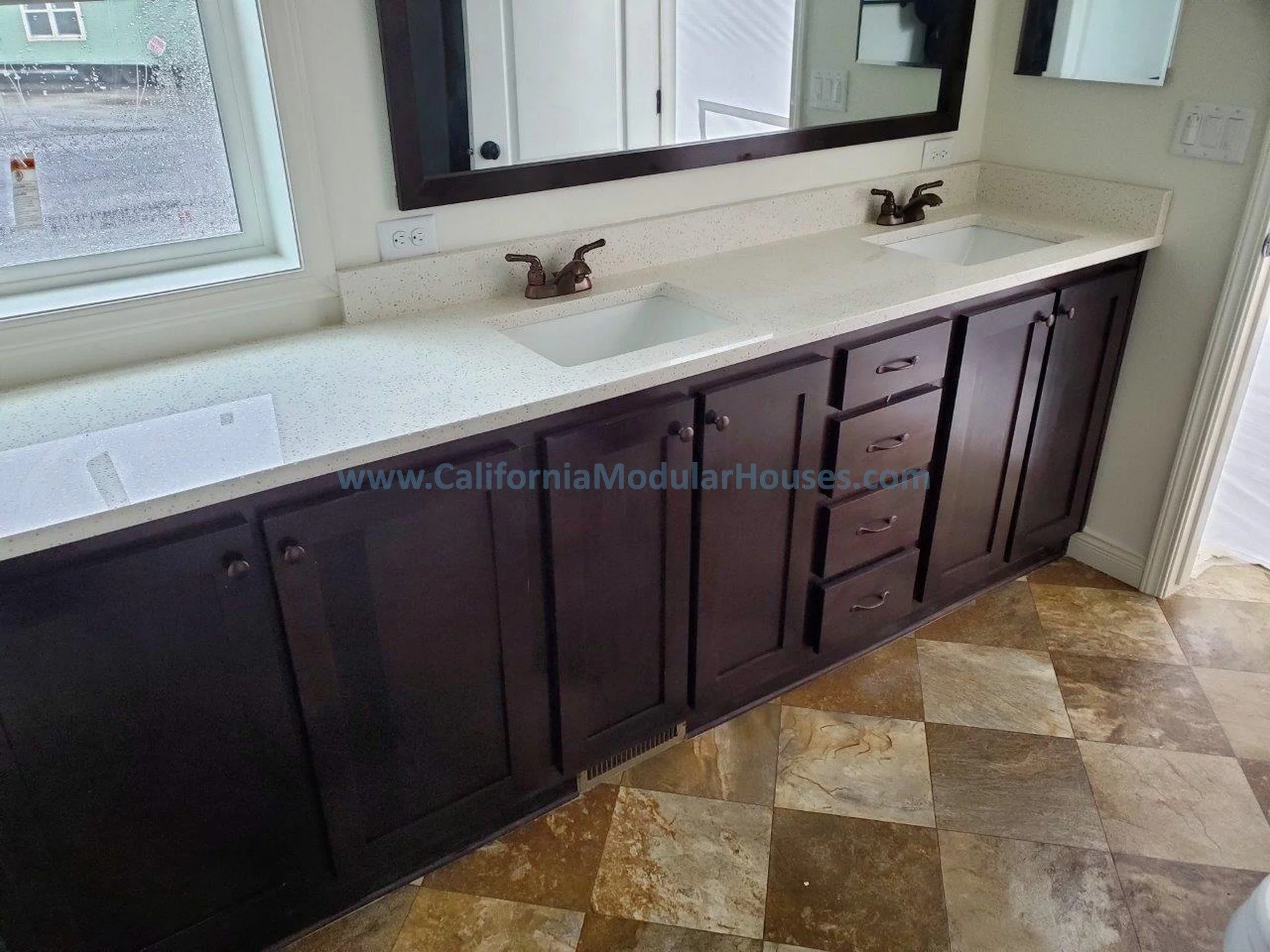 Double bathroom vanity with dark wood cabinets, white speckled countertop, and two sinks with matching bronze faucets, beside a window and a large rectangular mirror.   City of Santa Barbara, Santa Barbara County, CA.  Modular Home.  