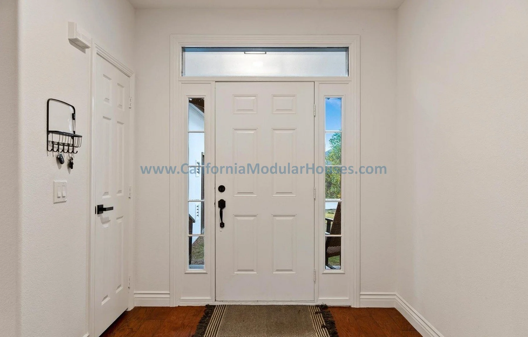 Entryway with a white front door, sidelights, and a large transom window above, leading outside to a small porch with trees visible in the background.
