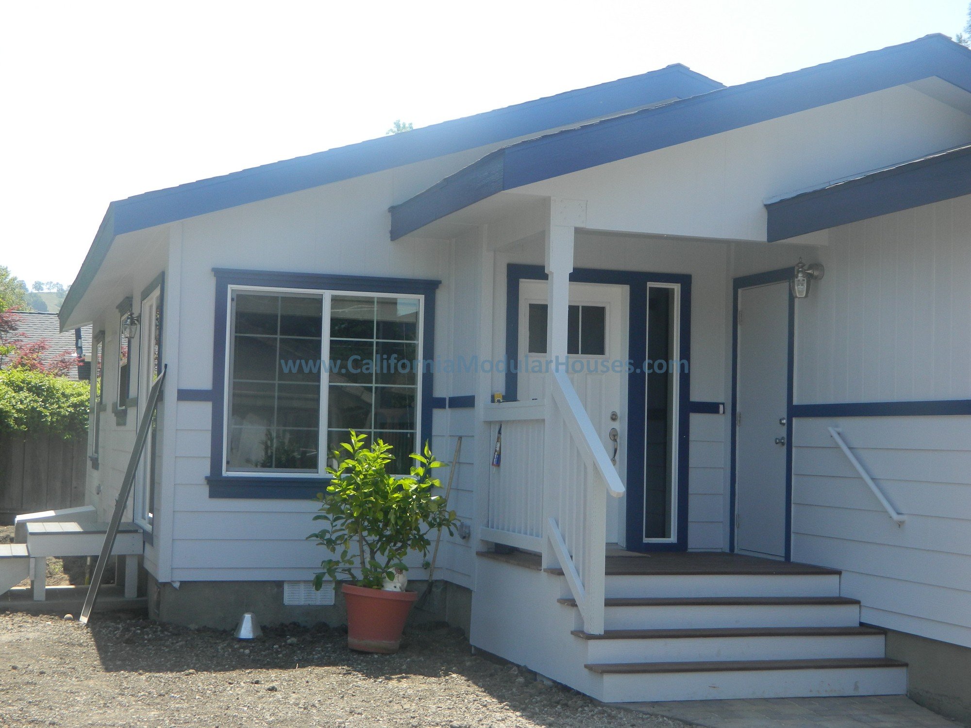 A small white house with blue trim, front porch, stairs, and a potted plant near the entrance, under a bright sky.