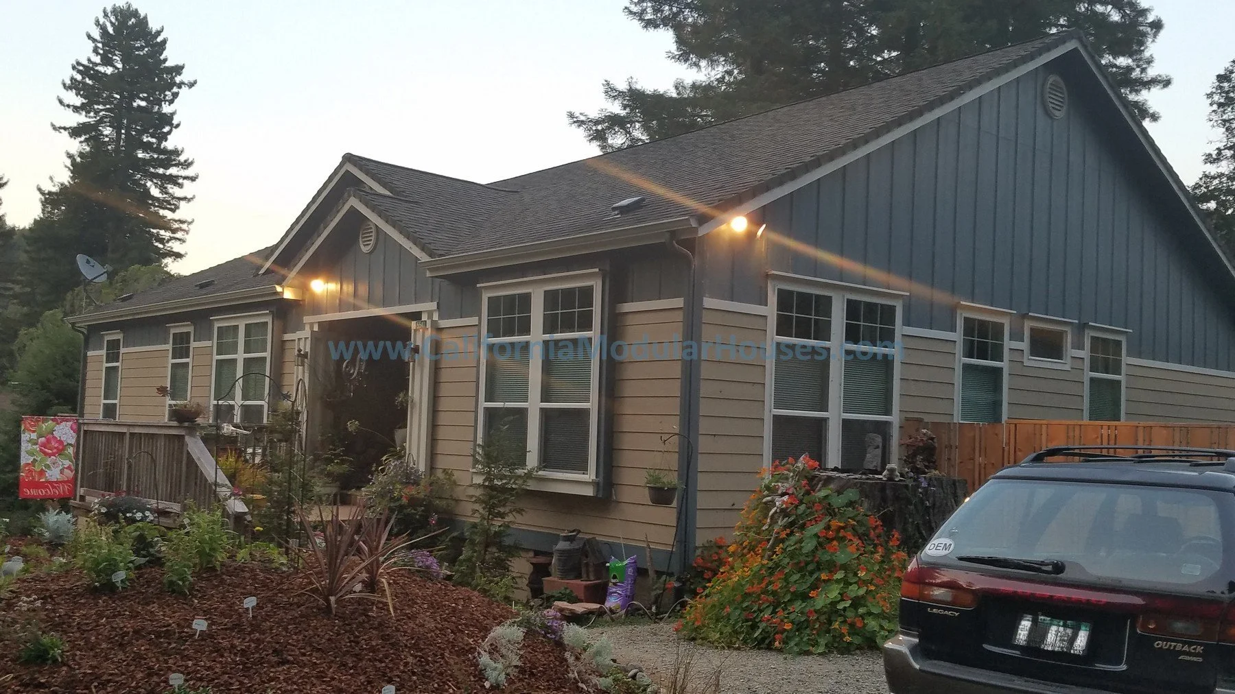 A two-story house with blue and beige siding, multiple windows, a garden in the front yard with plants and flowers, a driveway, and exterior lights turned on, surrounded by tall trees. Crescent City, Del Norte County, CA.  Modular Home.  