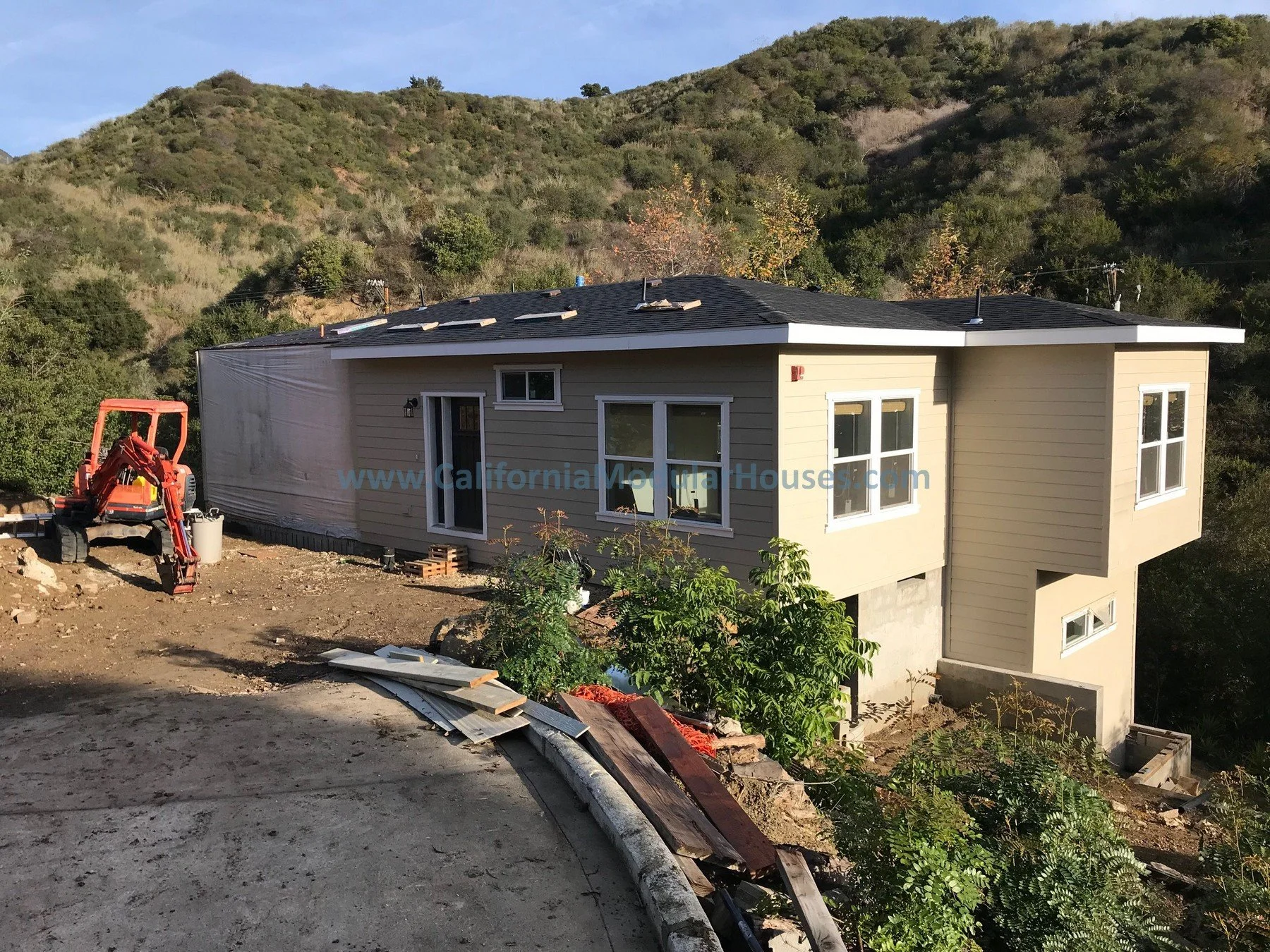 Construction site featuring a beige house with white trim on a hillside, with a small orange excavator on the left side, and trees and hills in the background.  City of Santa Barbara, Santa Barbara County, CA.  Modular Home.  