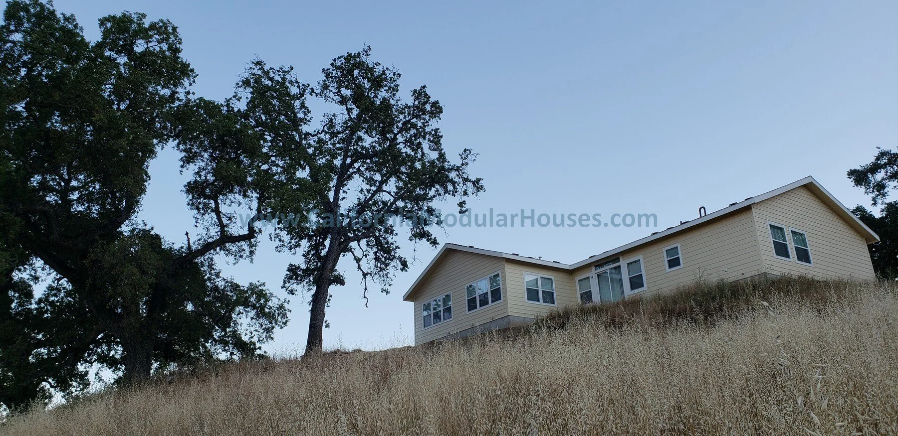 A beige modular house on a grassy hillside with a few large trees against a clear blue sky.  California Modular Houses, Modular Home CA.