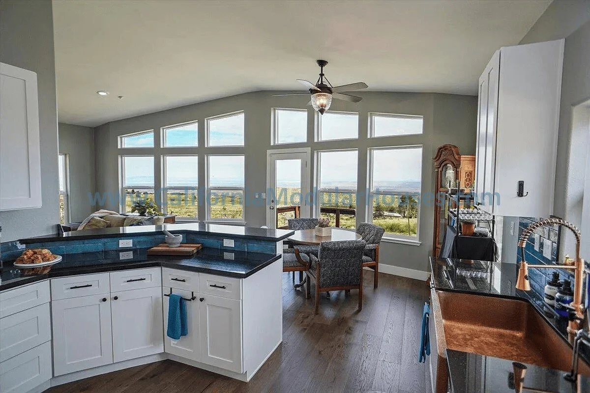 Kitchen with white cabinets, black countertops, and a copper sink, open to a dining area with a table and four chairs, large windows with a view of the outdoors, and a ceiling fan.