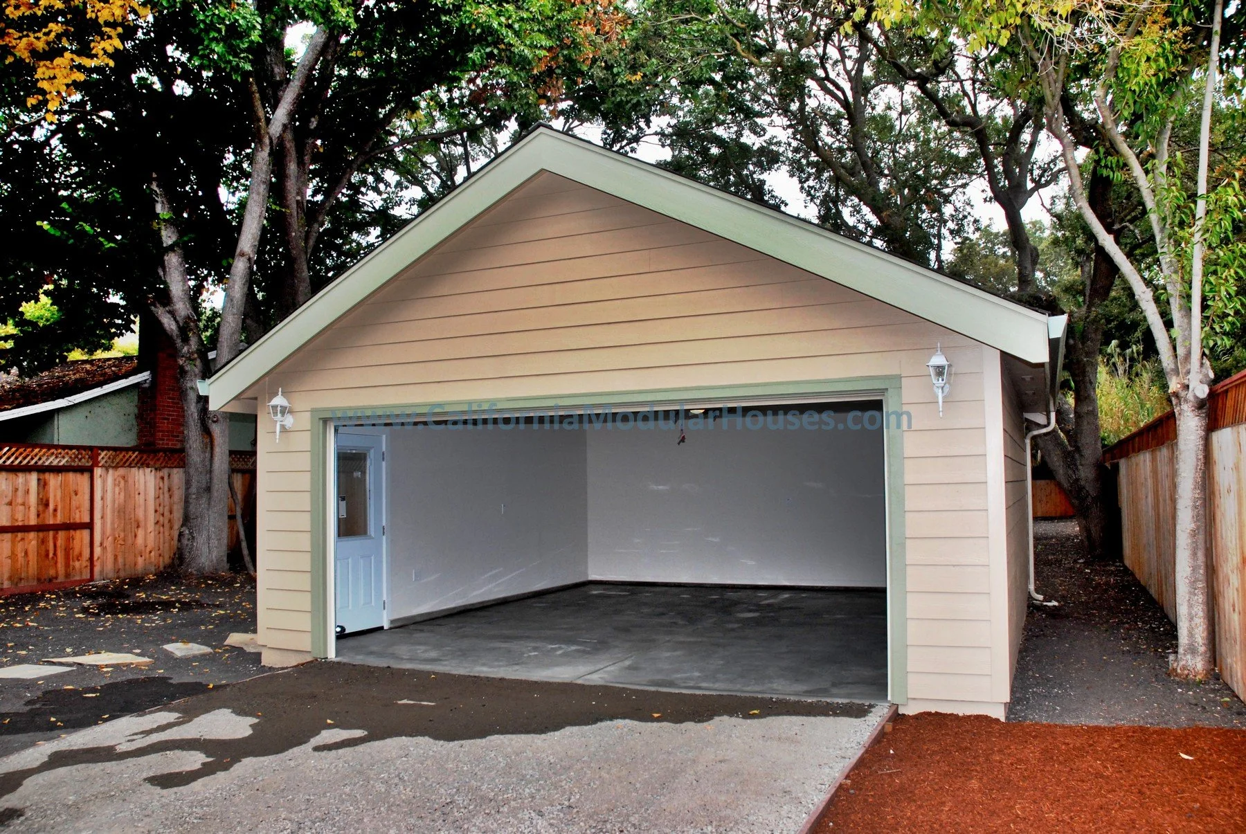 Newly built beige garage with open door, gray interior, concrete floor, surrounded by trees and wooden fences.