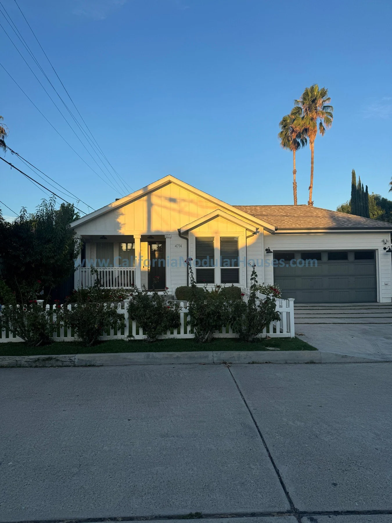 A white house with a gray roof and a white picket fence, surrounded by bushes, with a clear blue sky and tall palm trees in the background.