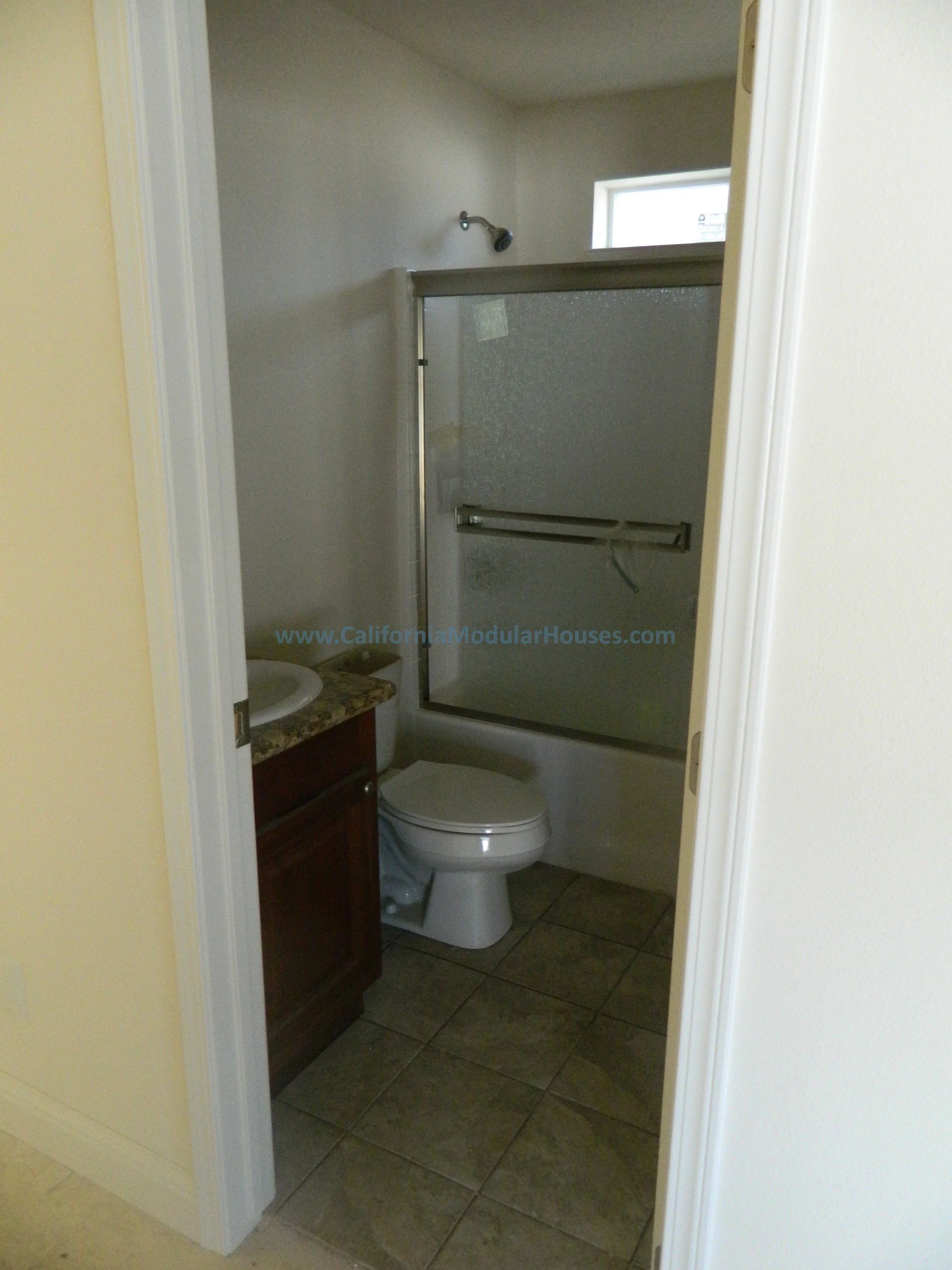 Small bathroom with a toilet, a sink with a dark wood cabinet, and a shower with a frosted glass door, beige tiled floor, white walls, a small window, and a shower head.