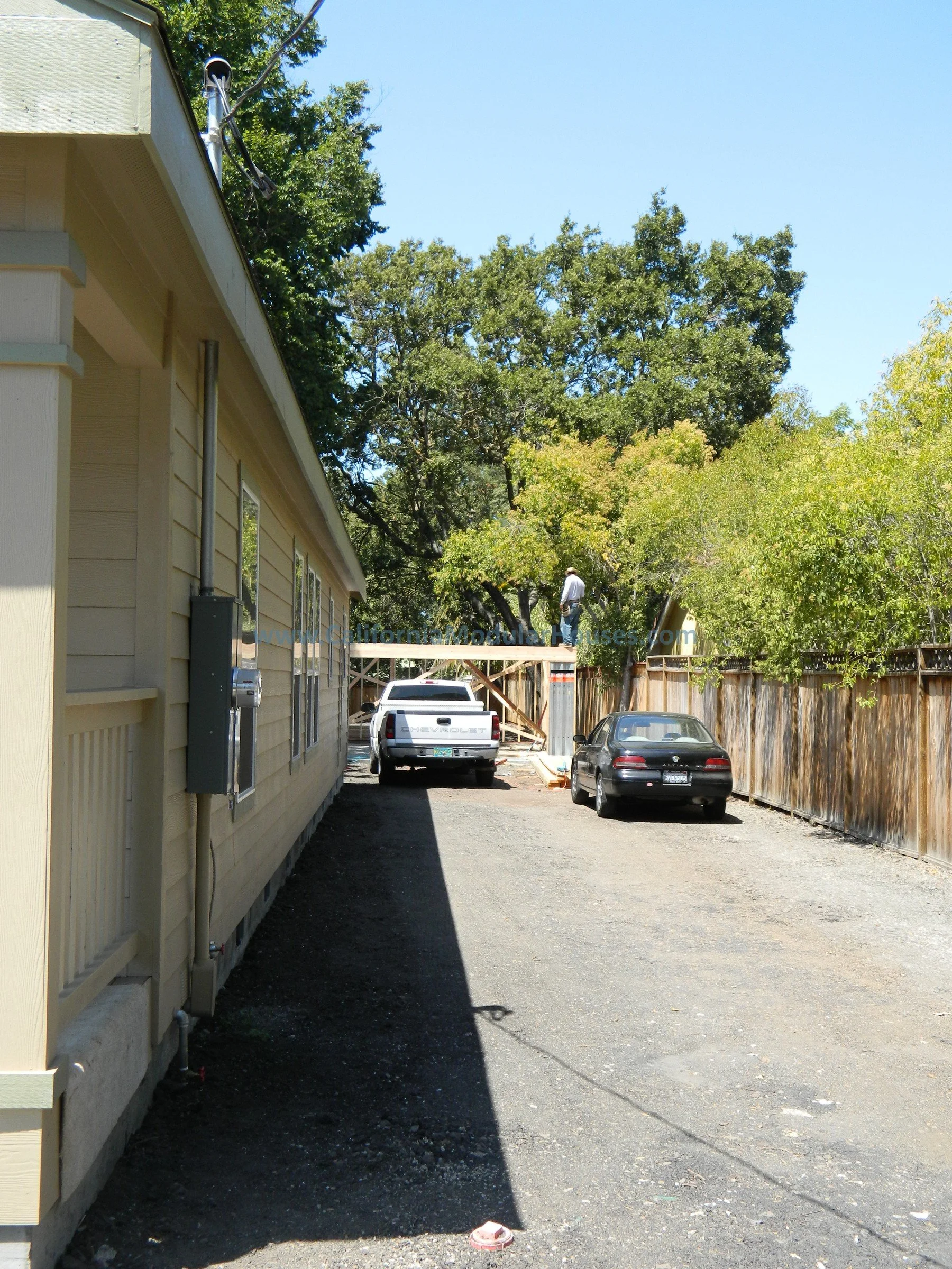 A narrow driveway beside a yellow house with electrical meters, with two parked cars and a person working on a wooden structure at the back, surrounded by trees and wooden fences.