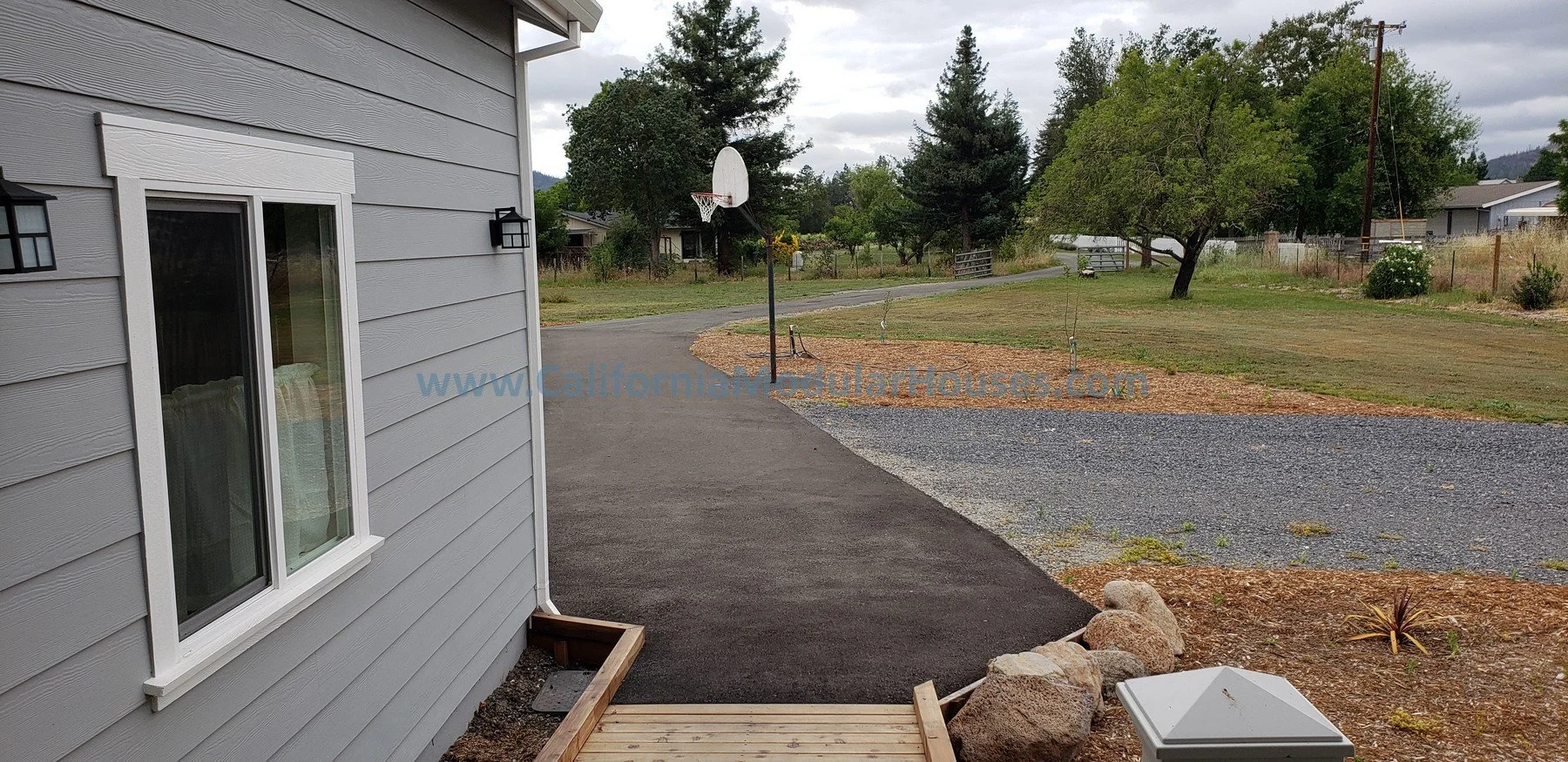 View of a suburban backyard with a gray house on the left, a gravel pathway, a basketball hoop, trees, and a cloudy sky.  2 bedroom, 2 bathroom modular ADU from the exterior.  Prefab Modular Accessory Dwelling Unit 