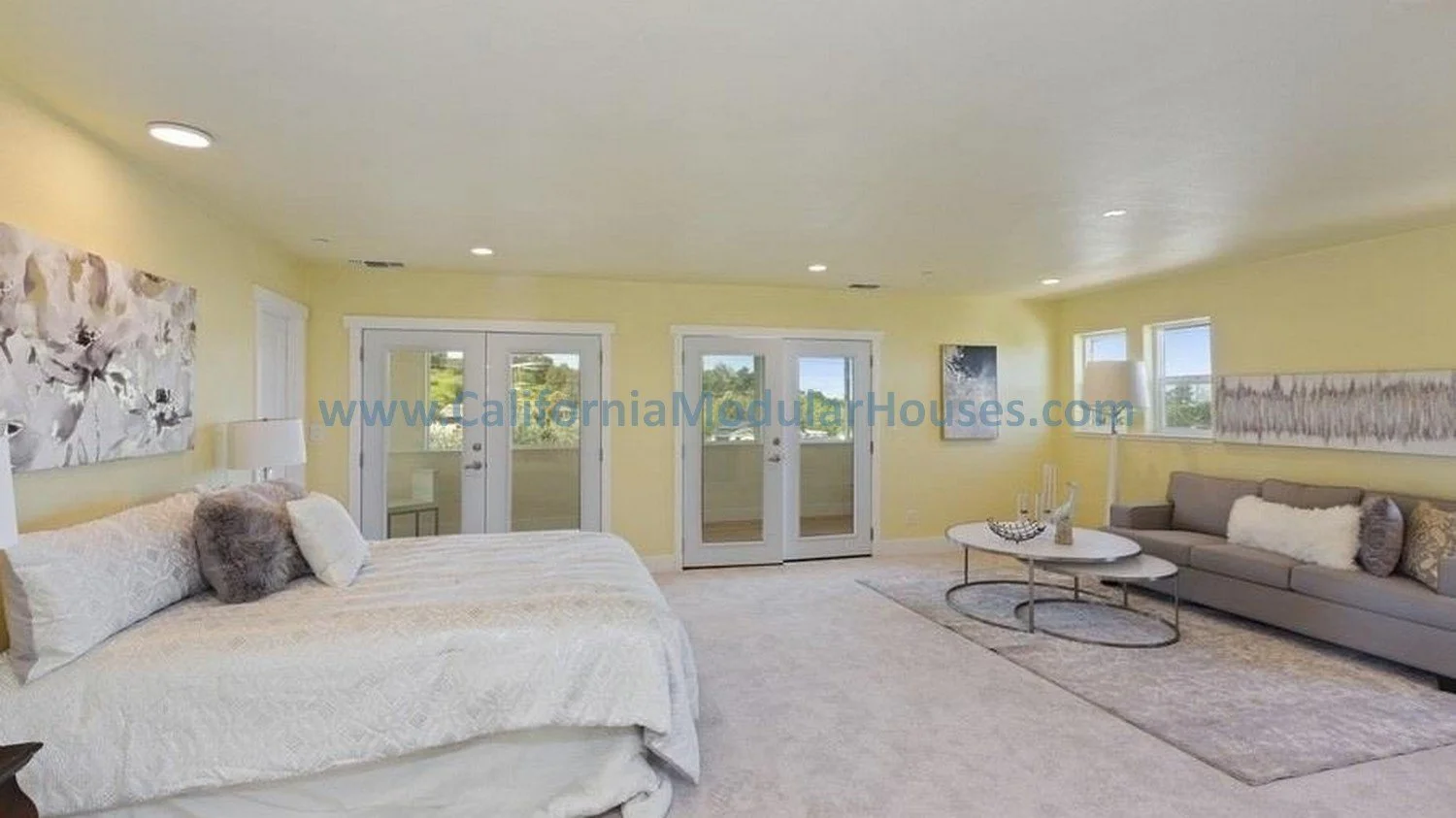 Living room with a bed on the left, a gray sofa on the right, and a glass door to a balcony in the center. Yellow walls, artwork, and windows adorn the room.  PreFab Modular for California. Contra Costa County 2 story modular home.  Modular house. 