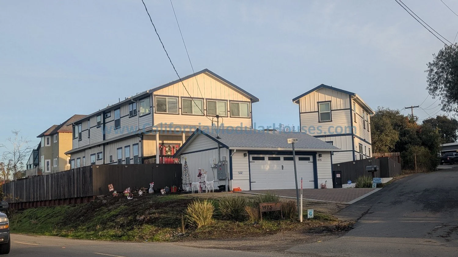 Multi-story house with a white exterior and a two-car garage, situated on a sloped street, decorated with Christmas figures in front, and visible street signs for Crestview.