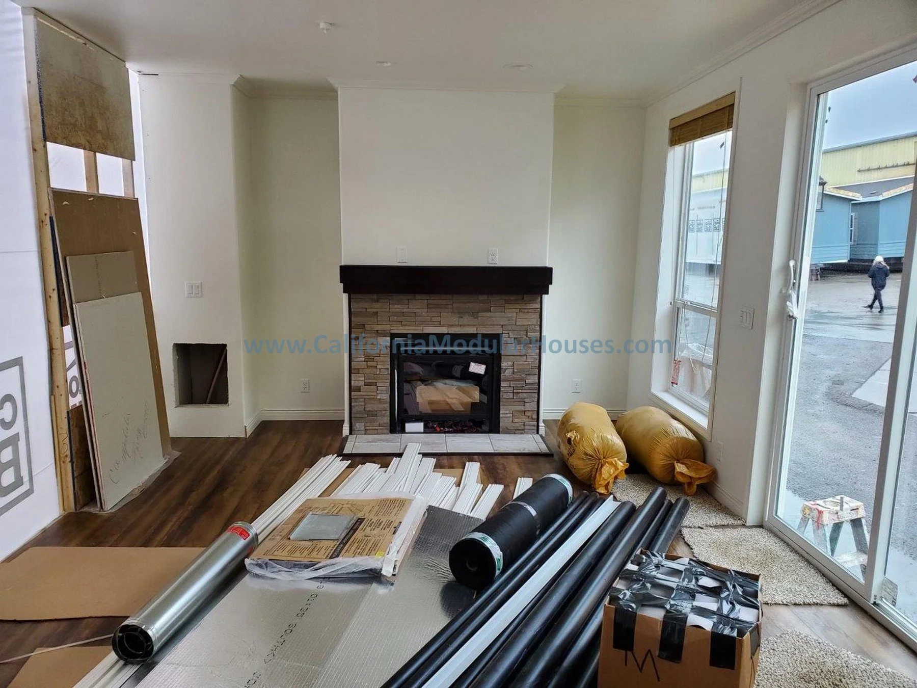 Living room under renovation with construction materials, pipes, and insulation on the floor, and a fireplace with a stone surround, flanked by large windows with blinds, and a sliding glass door leading outside.  Modular Homes.