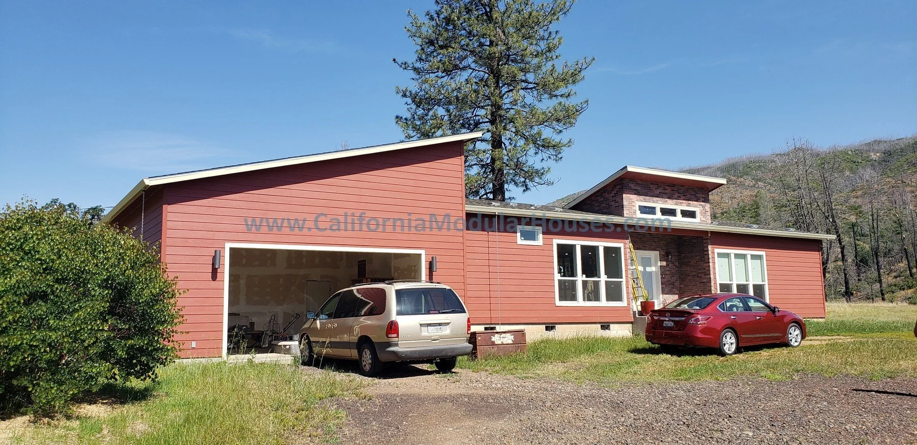 Modern two-story house with red siding, large windows, and garage.  The house is surrounded by green grass and trees with a mountain in the background under a clear blue sky. Prefab Modular Homes California, Prefab Modular Homes California.